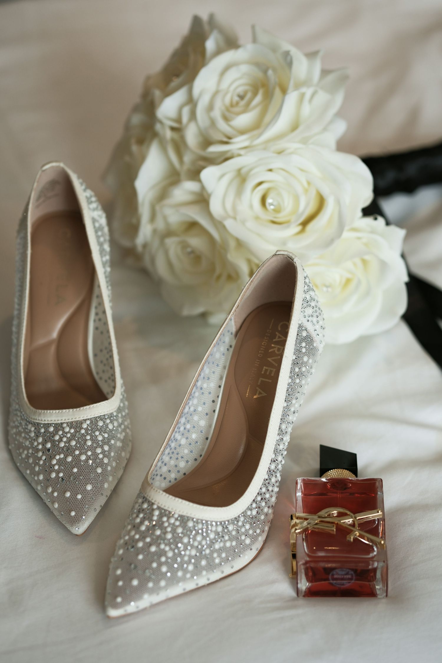 Sparkling crystal-embellished Carela bridal heels beside white rose bouquet and YSL Libre perfume bottle.