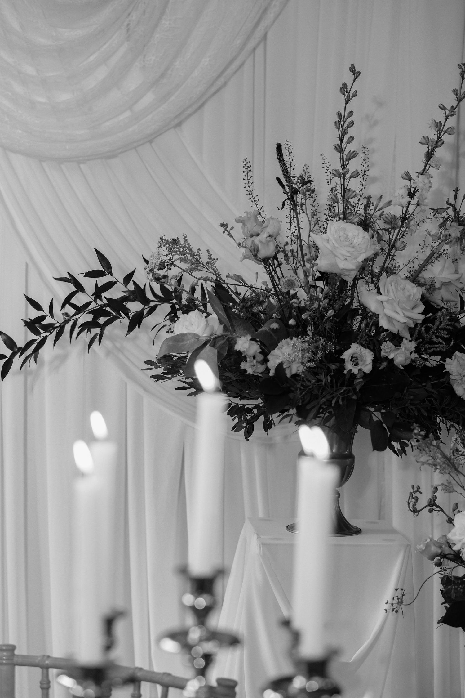 Black and white photo of elegant wedding floral arrangement with roses and candelabra against draped white backdrop.