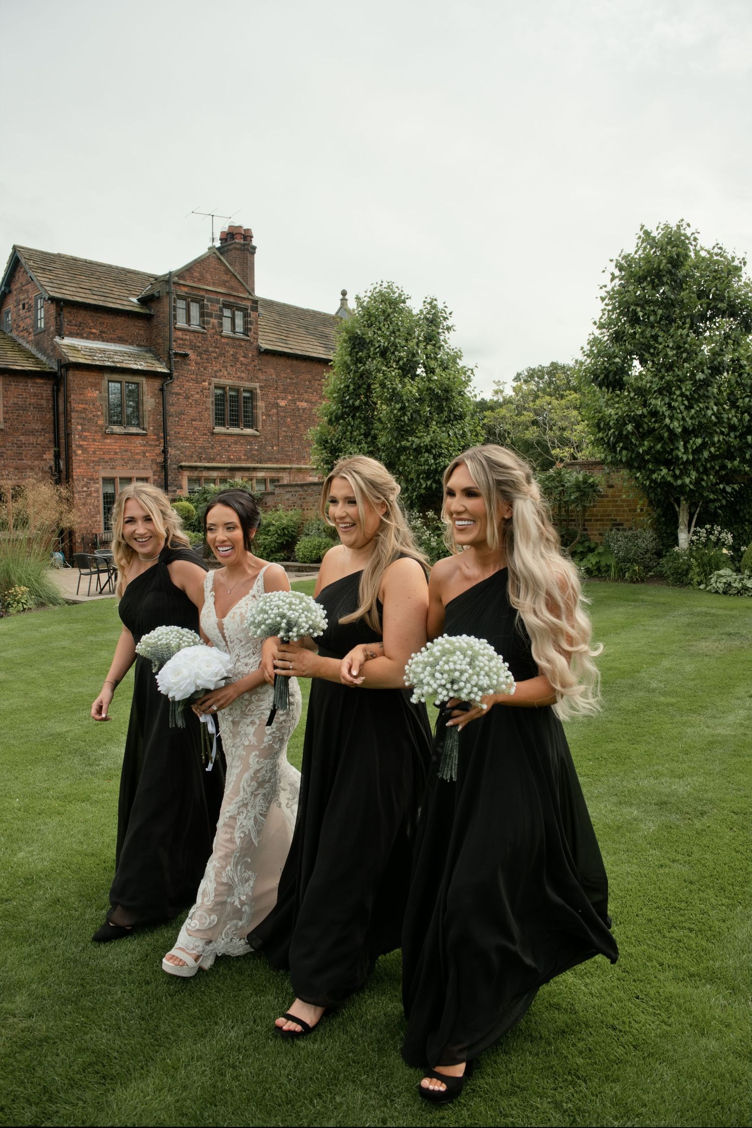 Bride in lace gown with three bridesmaids in black dresses holding blue hydrangea bouquets outdoors.