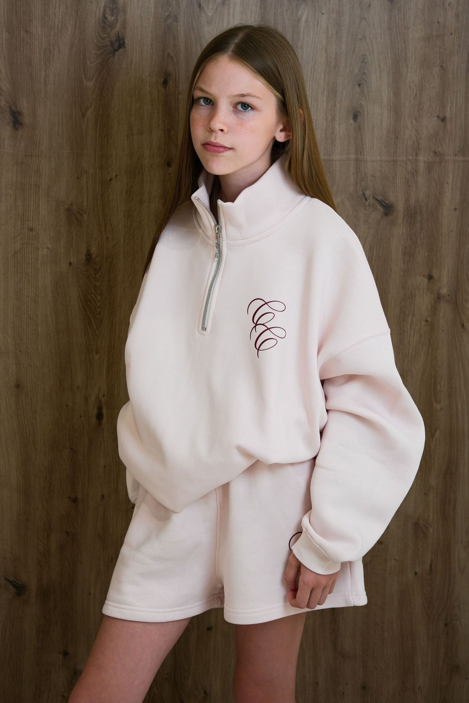 Young woman wearing an oversized light pink quarter-zip sweatshirt and matching shorts set with script logo embroidery.