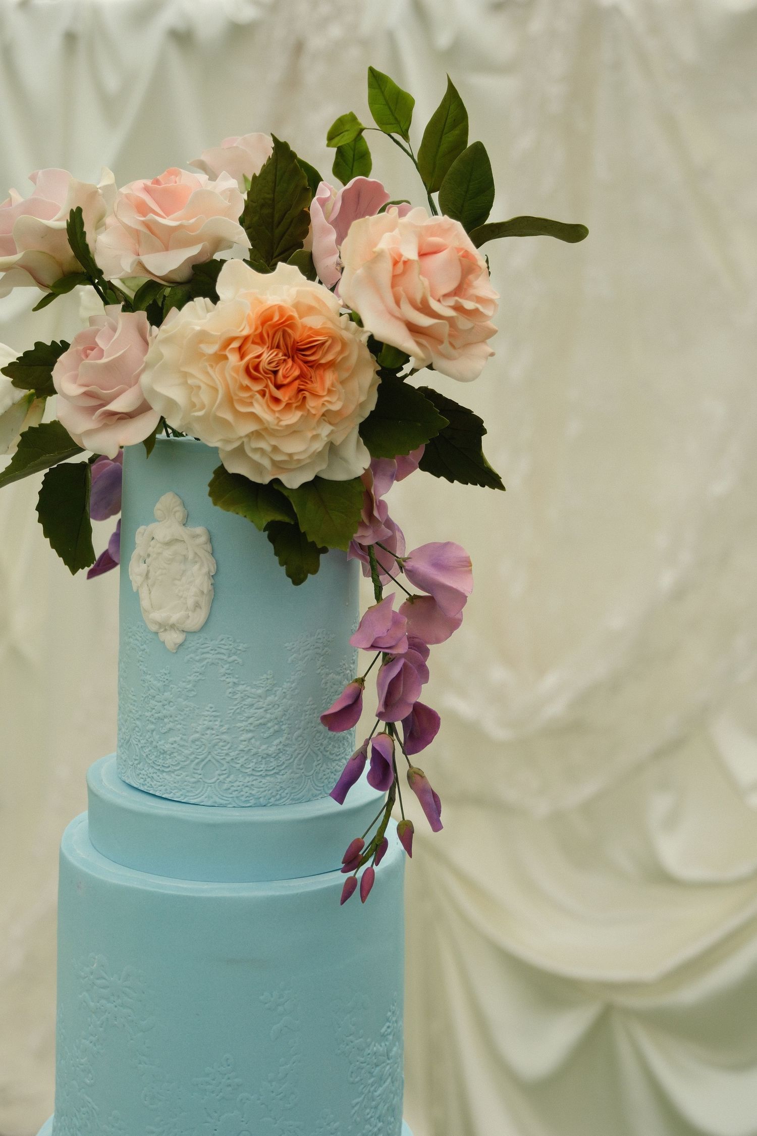 Elegant two-tier light blue wedding cake decorated with sugar roses and wisteria flowers.