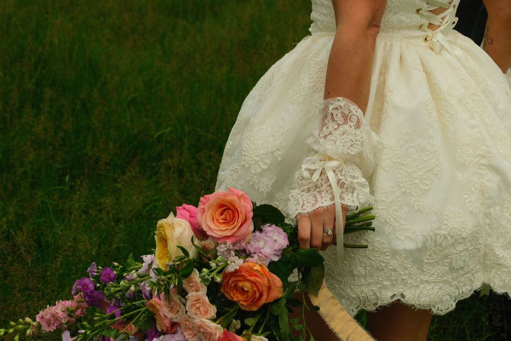 Bride in short lace wedding dress holding colorful bouquet of roses and wildflowers on green grass.