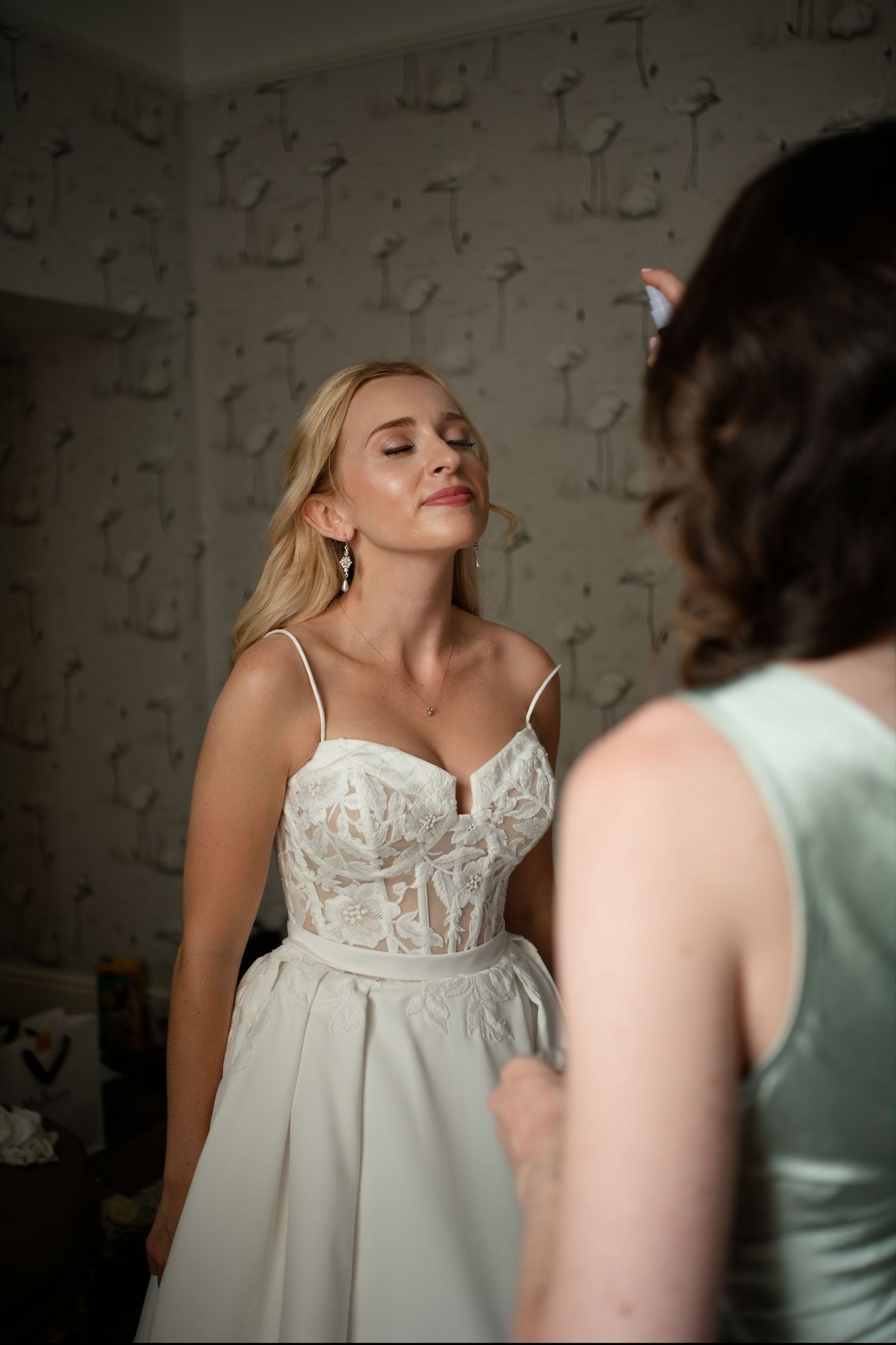 Blonde bride in lace wedding dress with eyes closed during bridal preparations in decorated room.