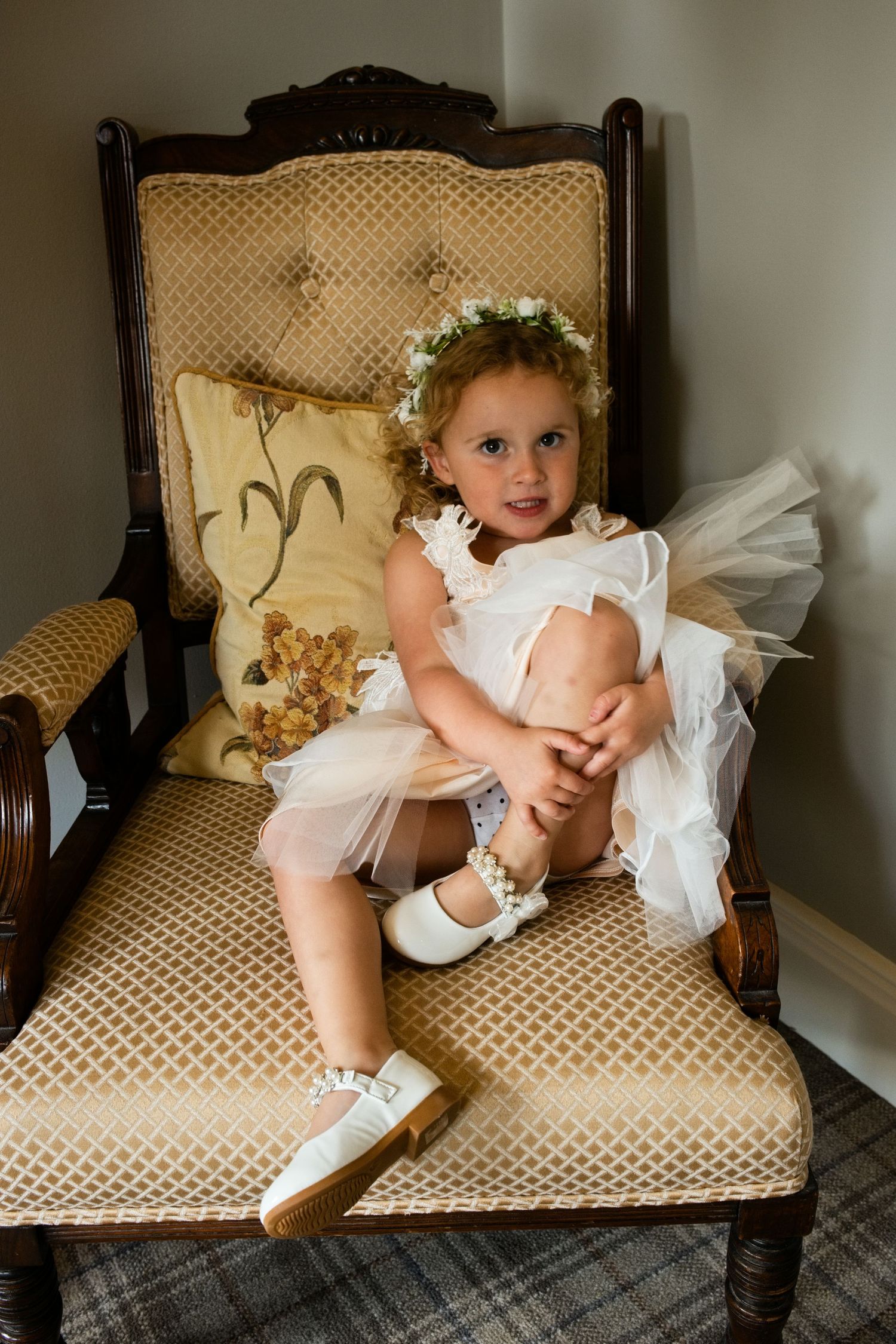 Adorable flower girl in white tutu dress with floral crown sitting on vintage gold armchair.