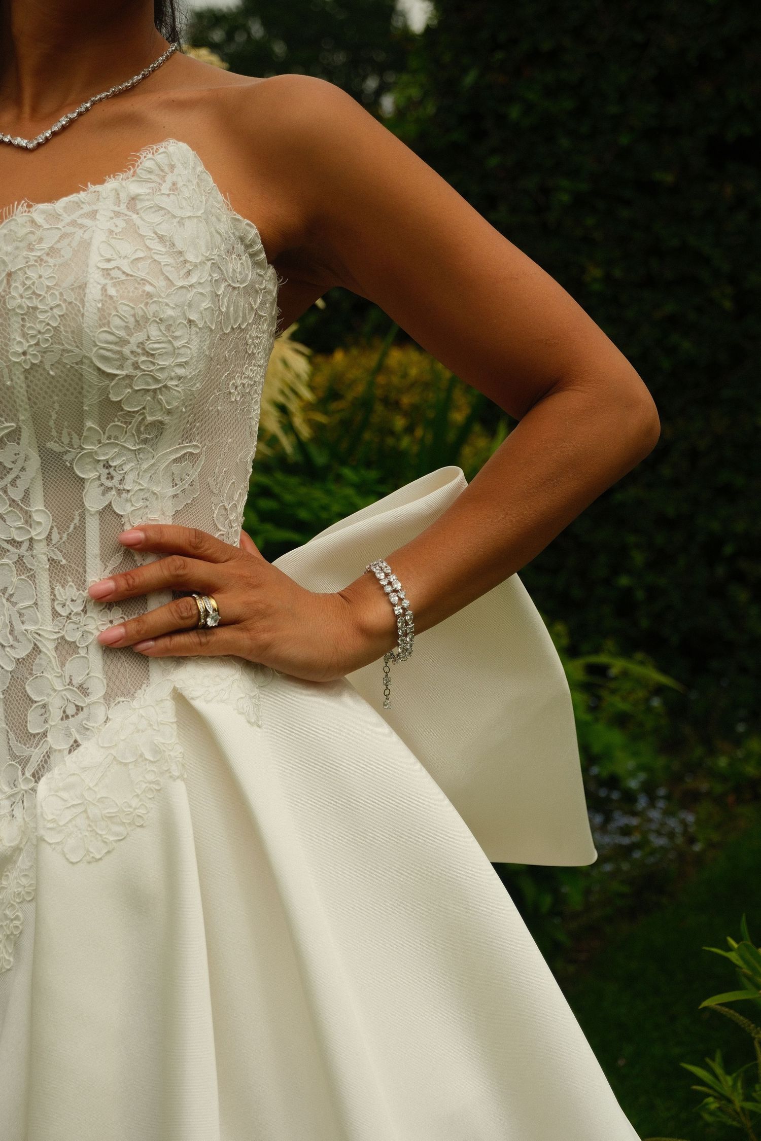 Close-up of bride in strapless lace corset wedding gown with large bow and pearl bracelet outdoors.