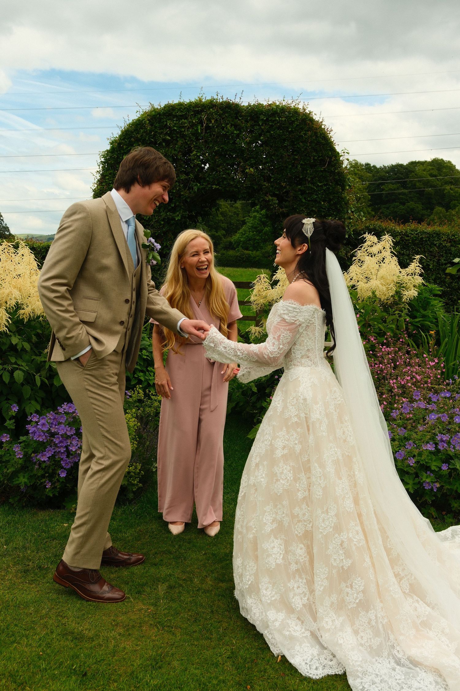 Bride in lace gown and groom in tan suit sharing a moment with bridesmaid in pink in garden.