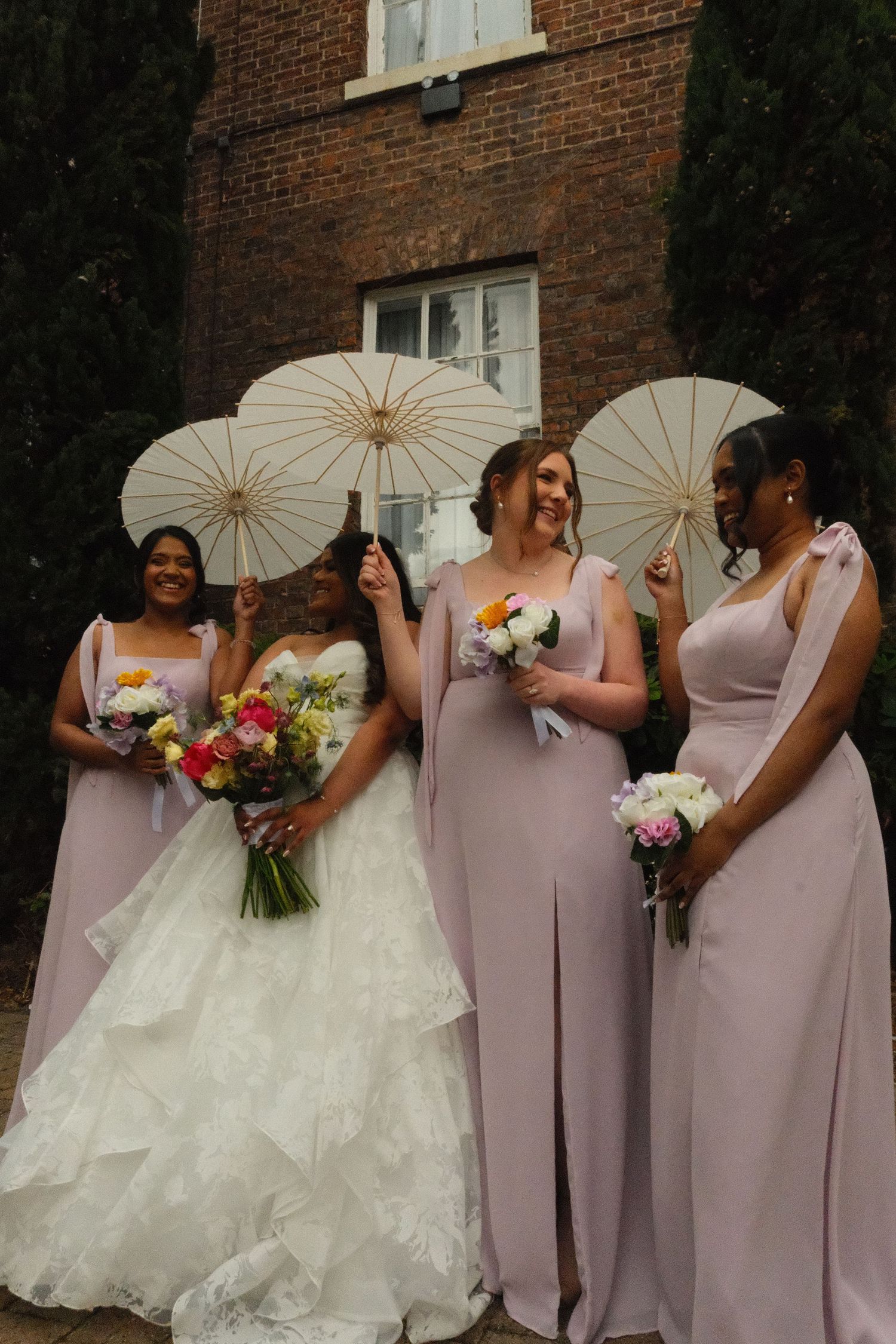 Bride in white gown with three bridesmaids in dusty pink dresses holding white parasols outside a brick building.