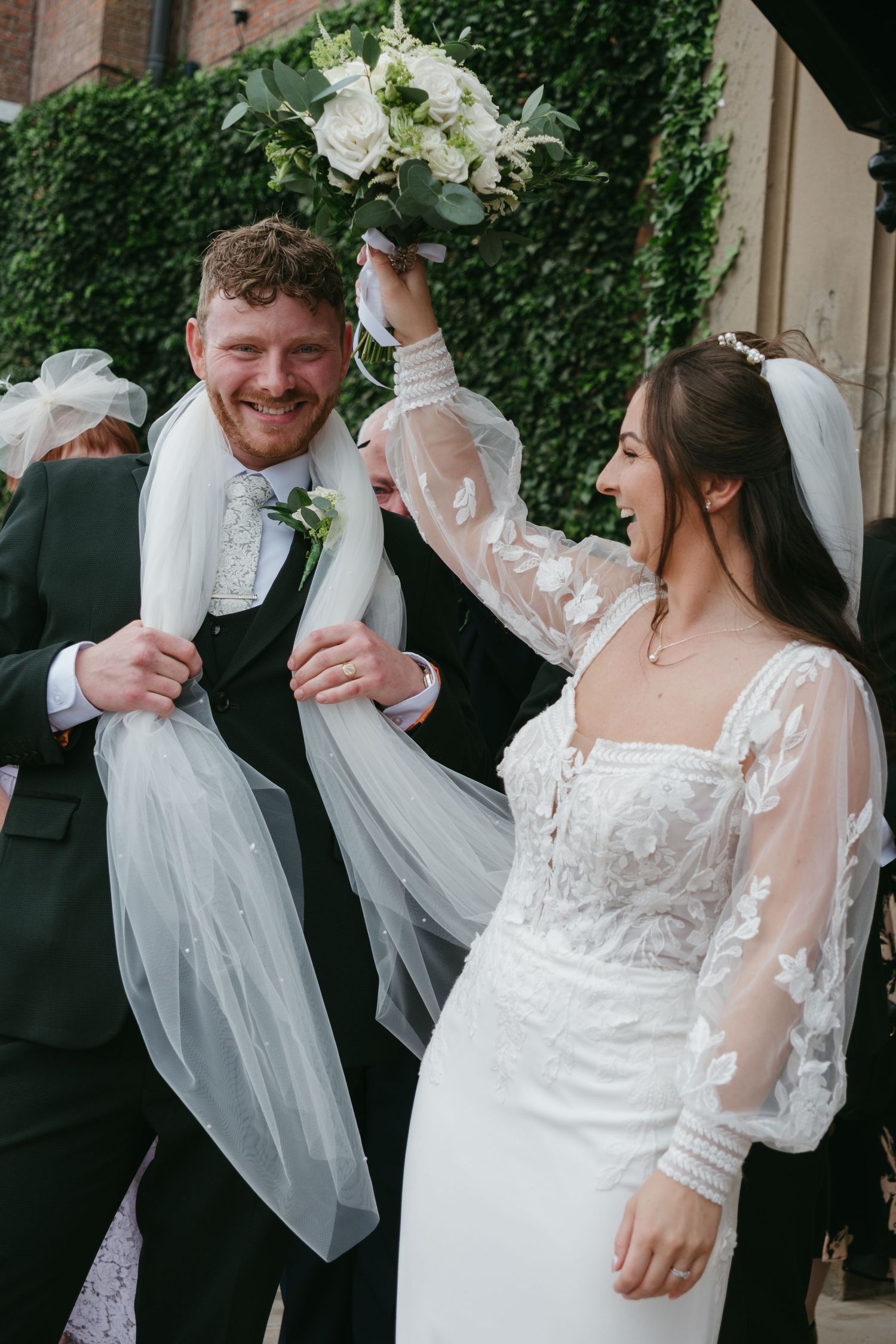 Joyful newlyweds celebrate outside church, bride raising bouquet while groom laughs with veil draped around him.