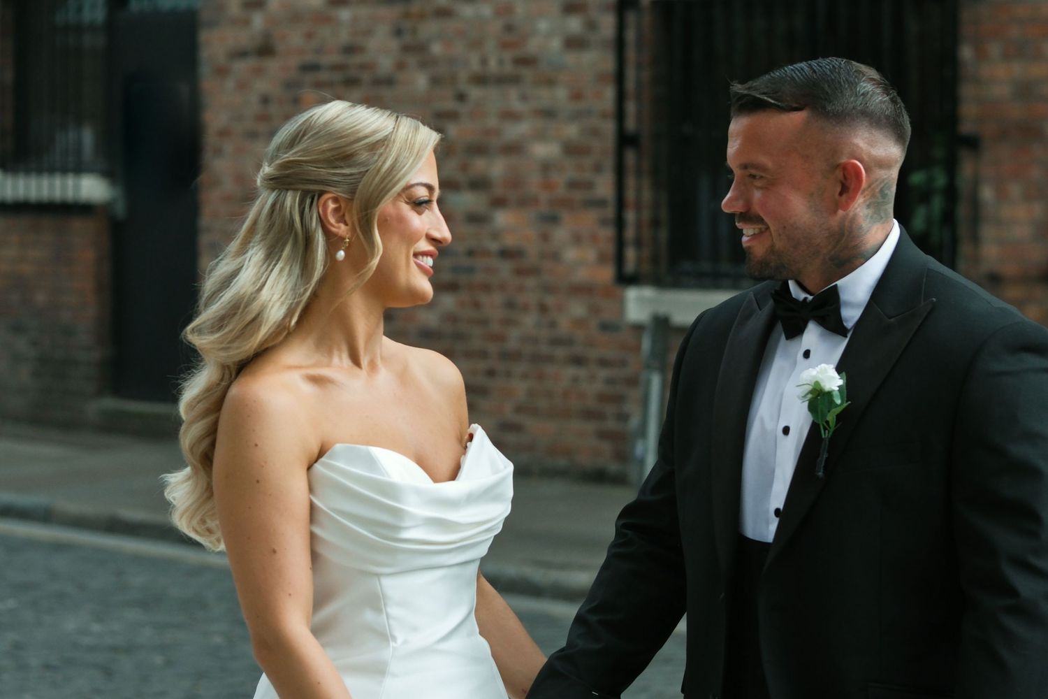 Smiling bride and groom holding hands facing each other on cobblestone street with brick wall backdrop.