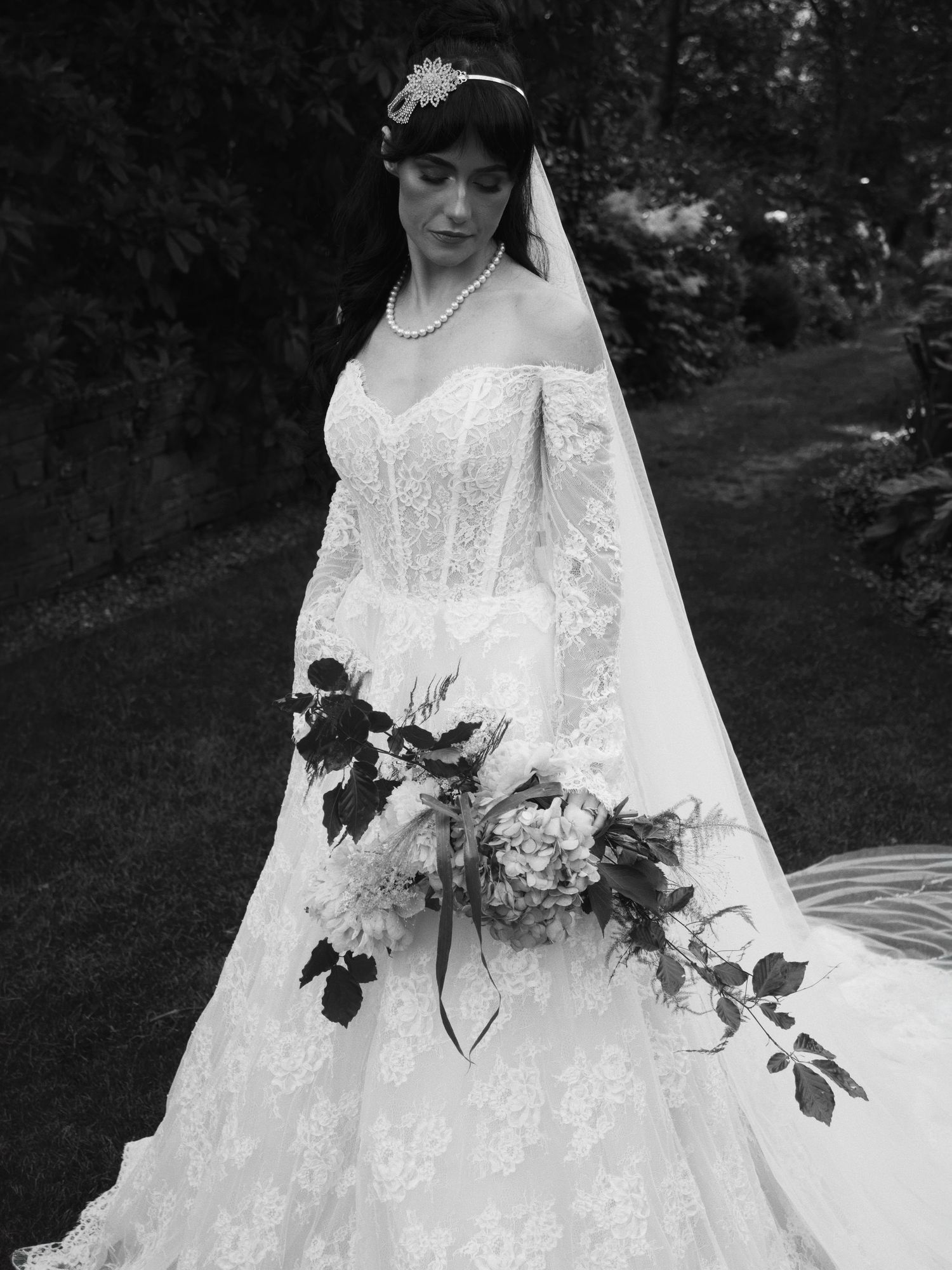 Black and white photo of a bride in lace off-shoulder gown holding floral bouquet outdoors.