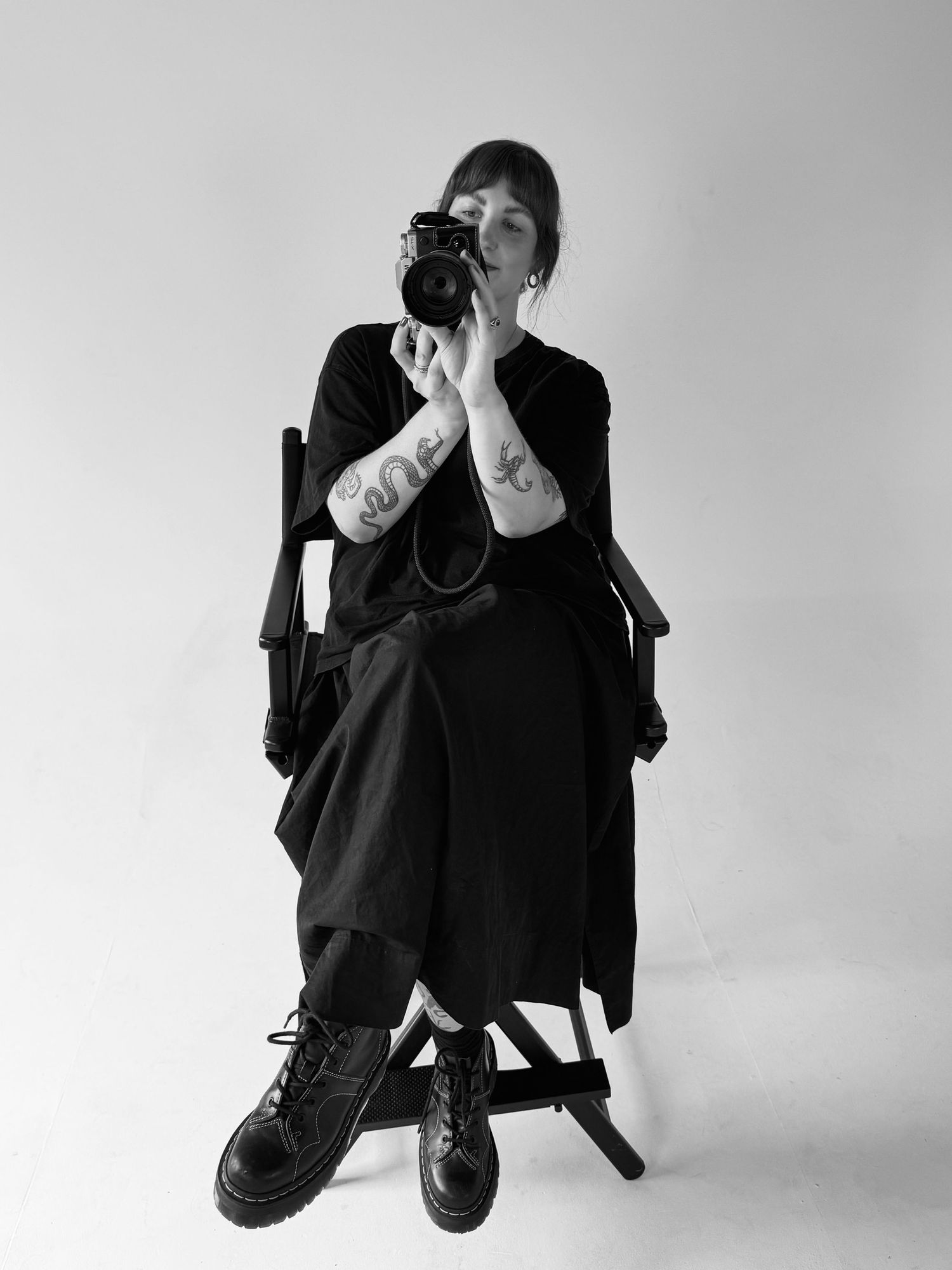 Black and white photo of a tattooed woman in black dress sitting in director chair holding a camera.