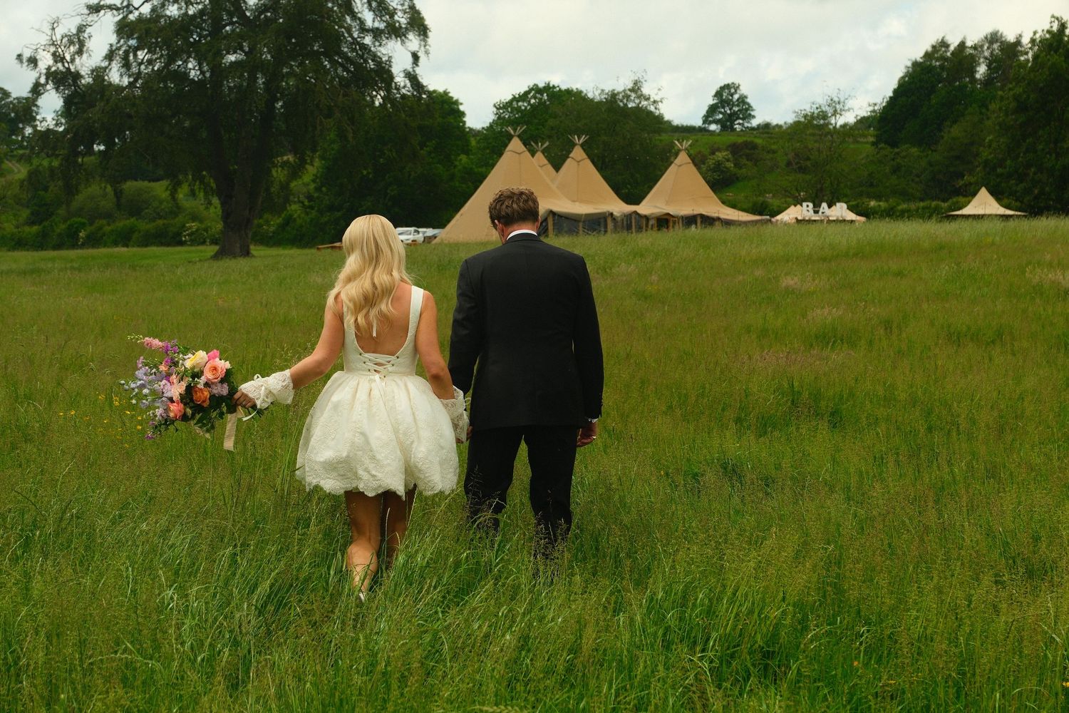 Bride and groom walk hand in hand through green meadow toward tipi wedding venue.