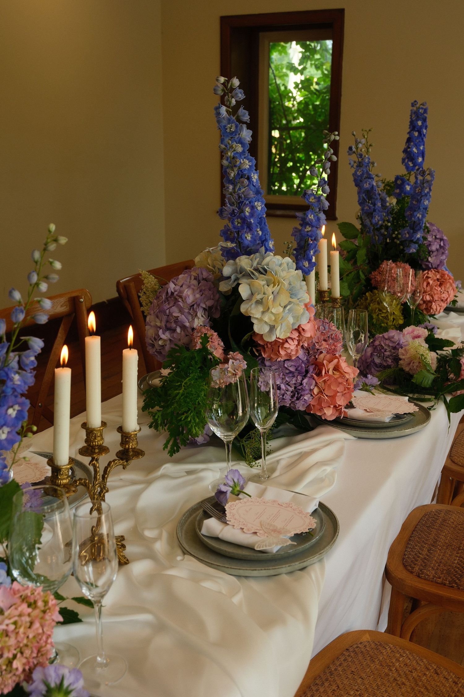 Elegant wedding reception table with white linen, candles, and colorful floral centerpieces of blue delphiniums and pink hydrangeas.