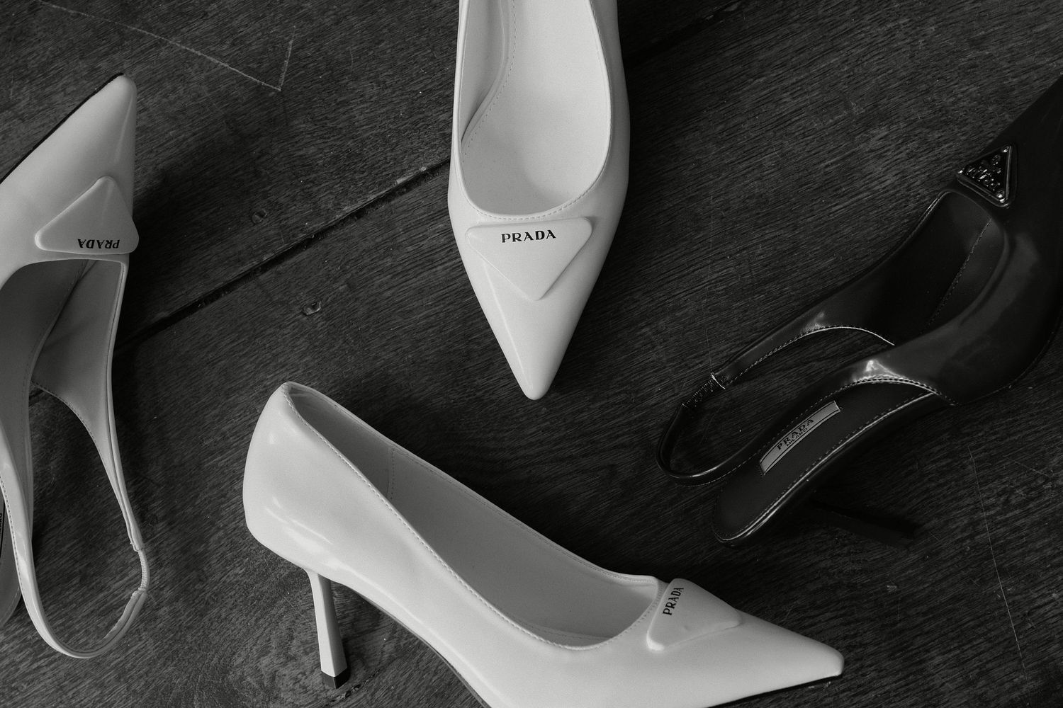 Black and white photo of Prada branded white pointed-toe heels and black strappy pumps arranged on a wooden floor.