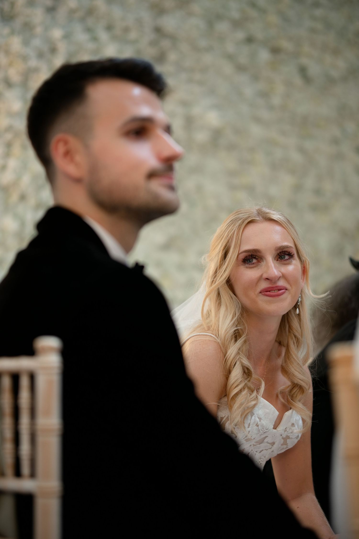 Bride in white lace dress gazes lovingly at groom in black suit during wedding ceremony.
