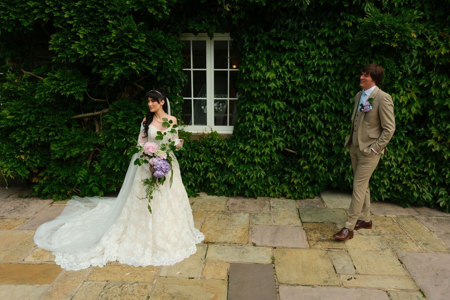 Bride in lace wedding gown with cascading bouquet and groom in tan suit beside ivy-covered wall.