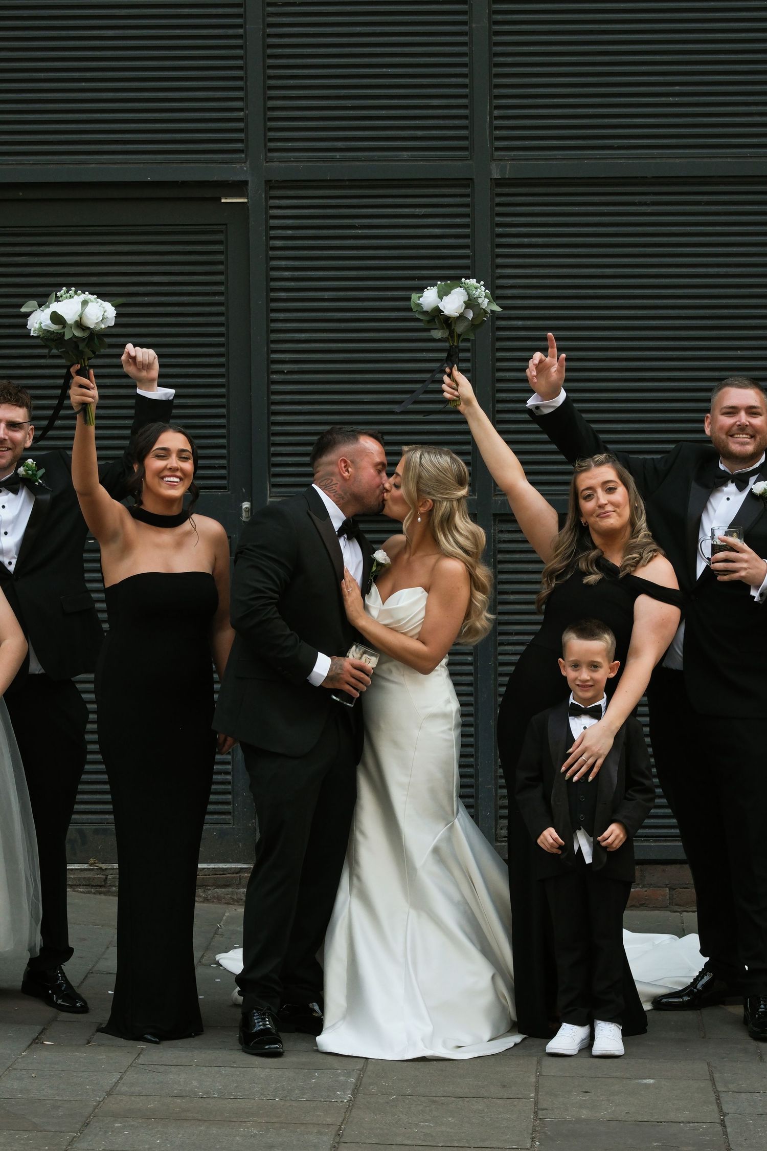 Wedding party celebrates as bride and groom kiss, bridesmaids and groomsmen in black cheer with bouquets raised.