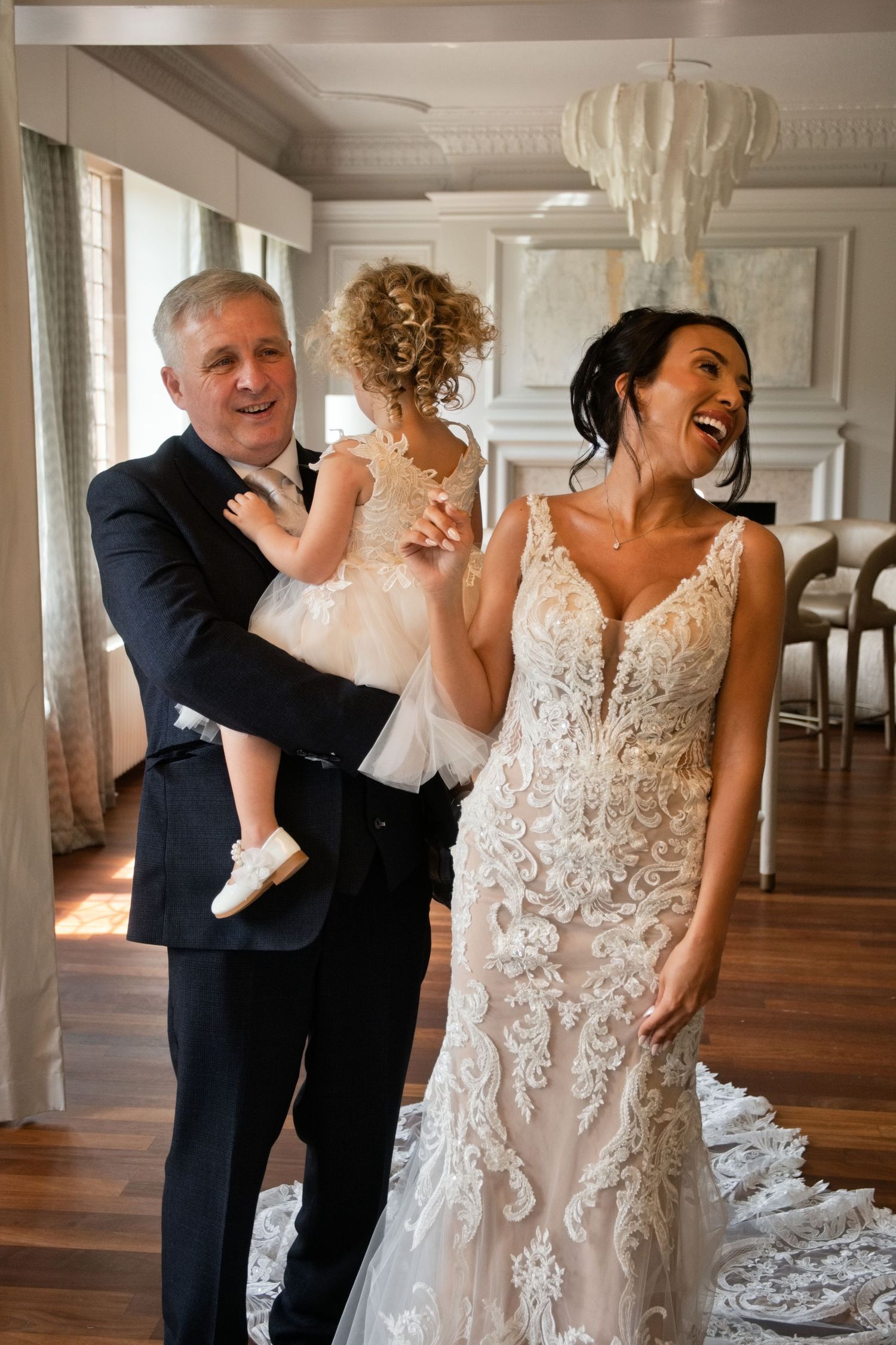 Laughing bride in lace wedding gown stands with older man holding curly-haired flower girl in white dress.