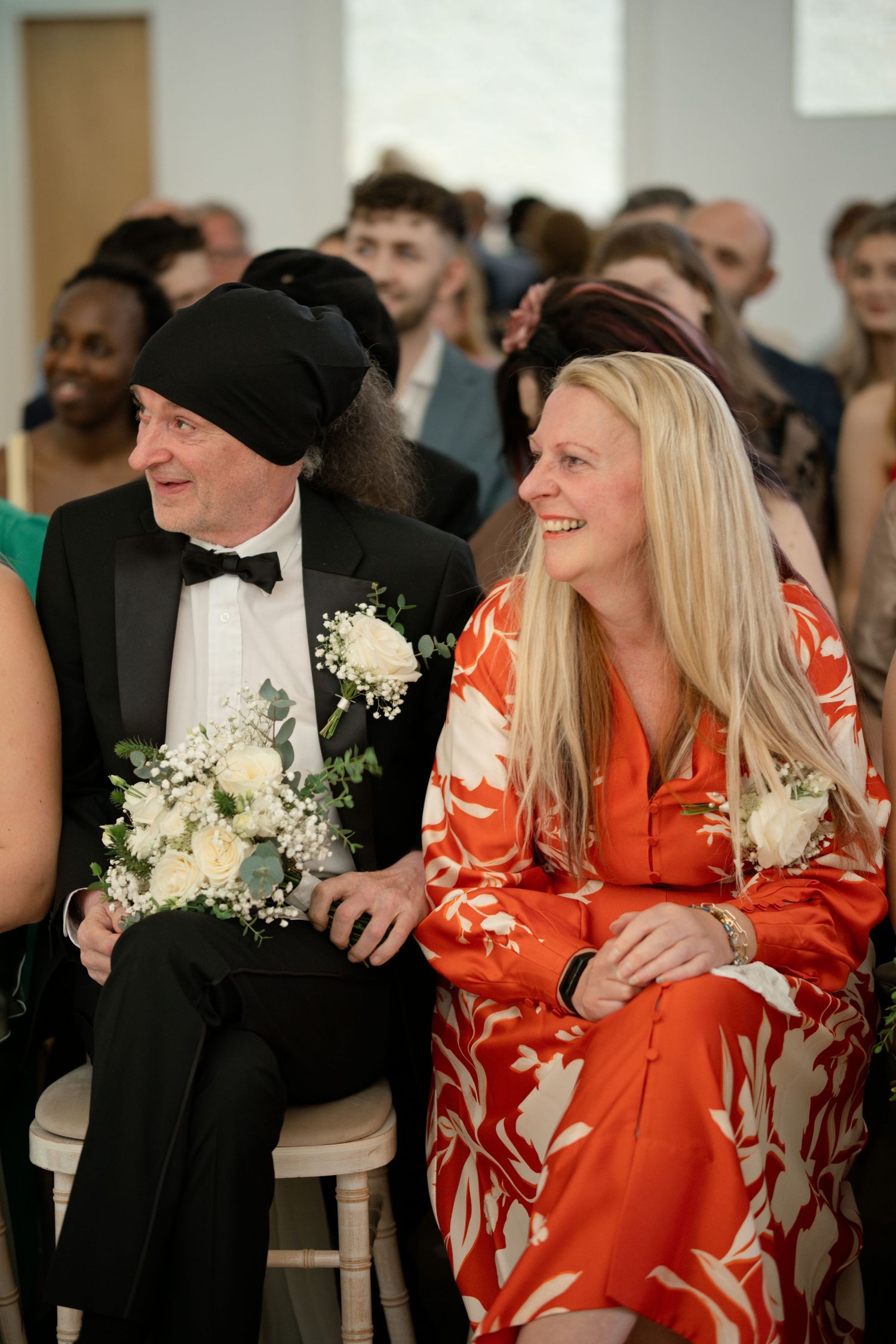 Wedding guests smiling and laughing during ceremony, man in black tux holding white bouquet.