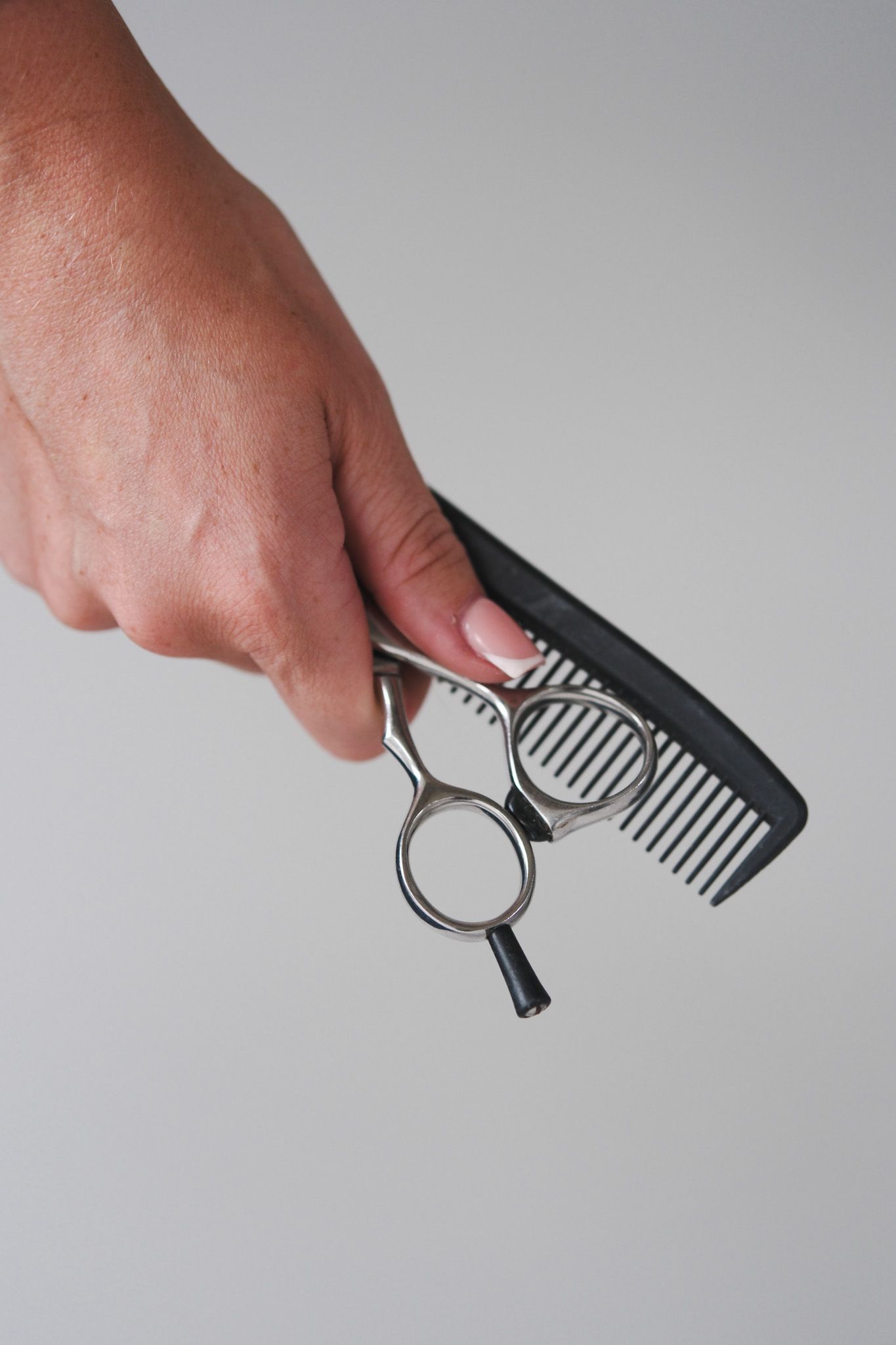 Hand holding professional hair cutting scissors and a black comb against a light gray background.