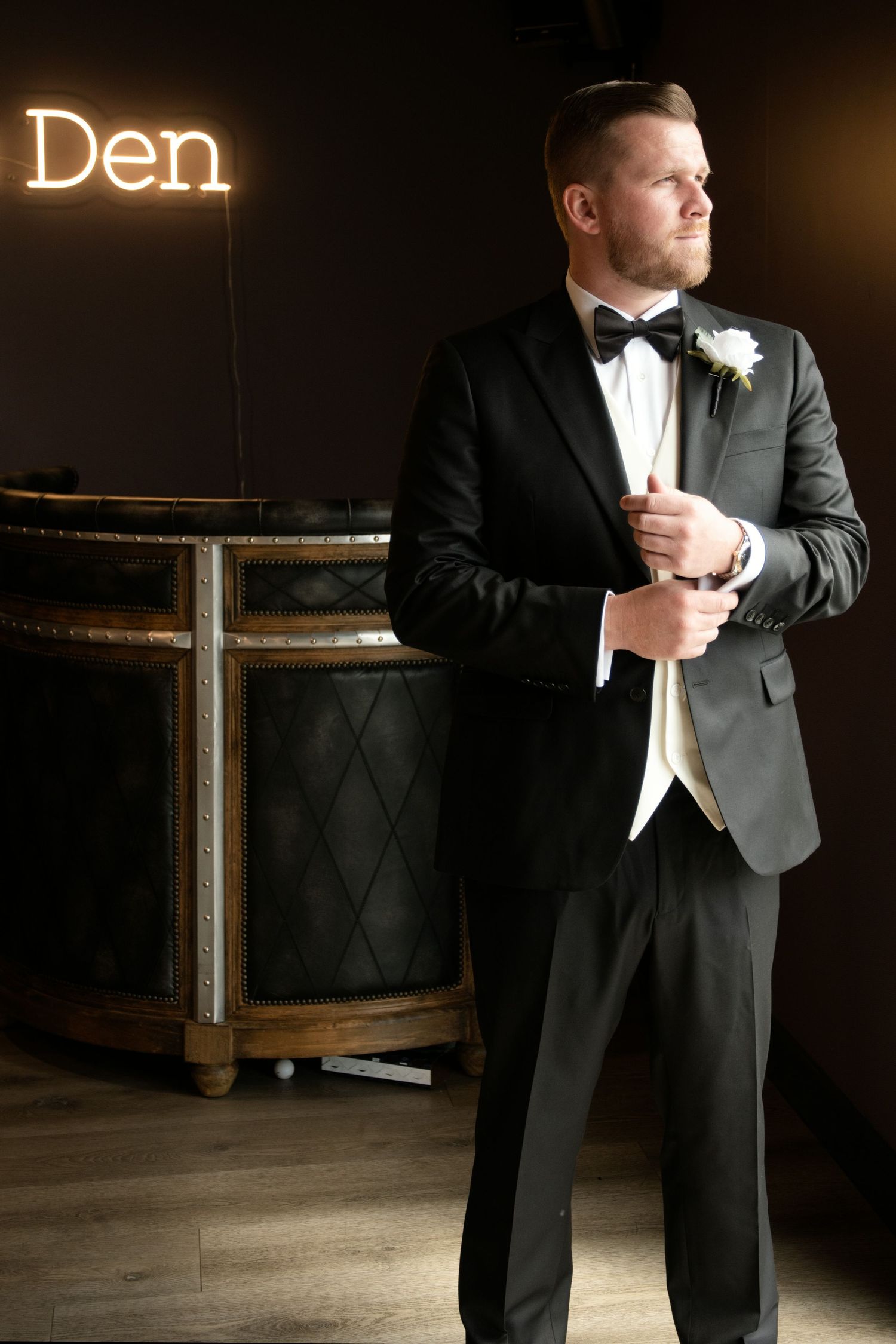 Dapper groom in black tuxedo with bow tie and boutonniere adjusts cufflinks beside glowing Den neon sign.