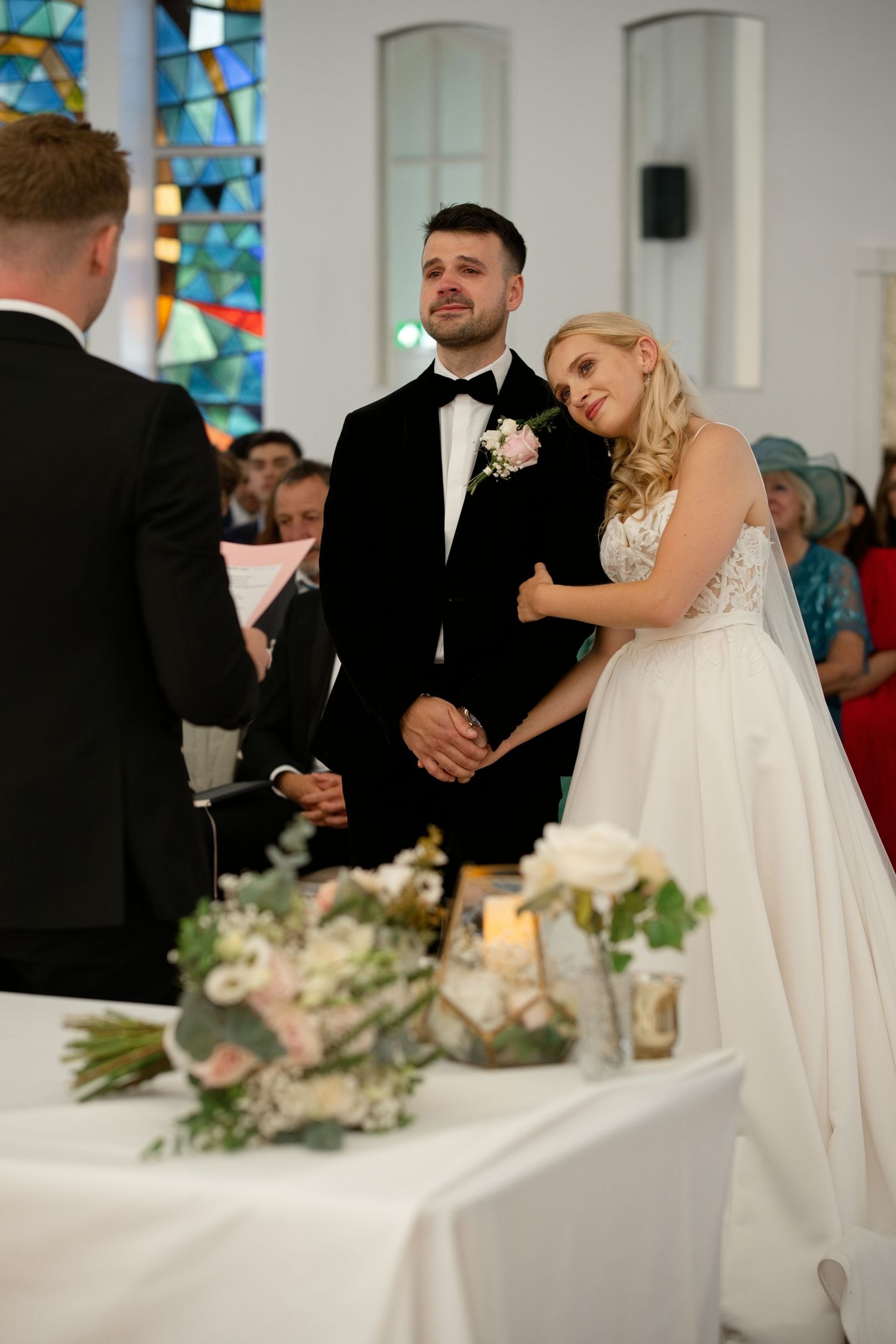 Bride and groom smiling at officiant during wedding ceremony with floral arrangements on altar.