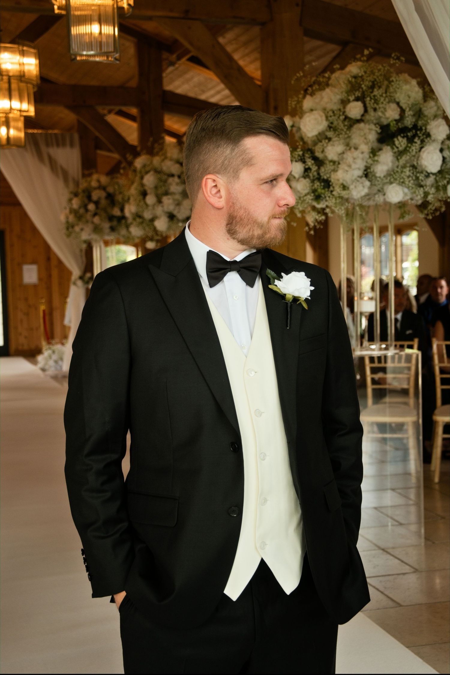Dapper groom in black tuxedo with bow tie and white boutonniere stands in floral-decorated wedding venue.