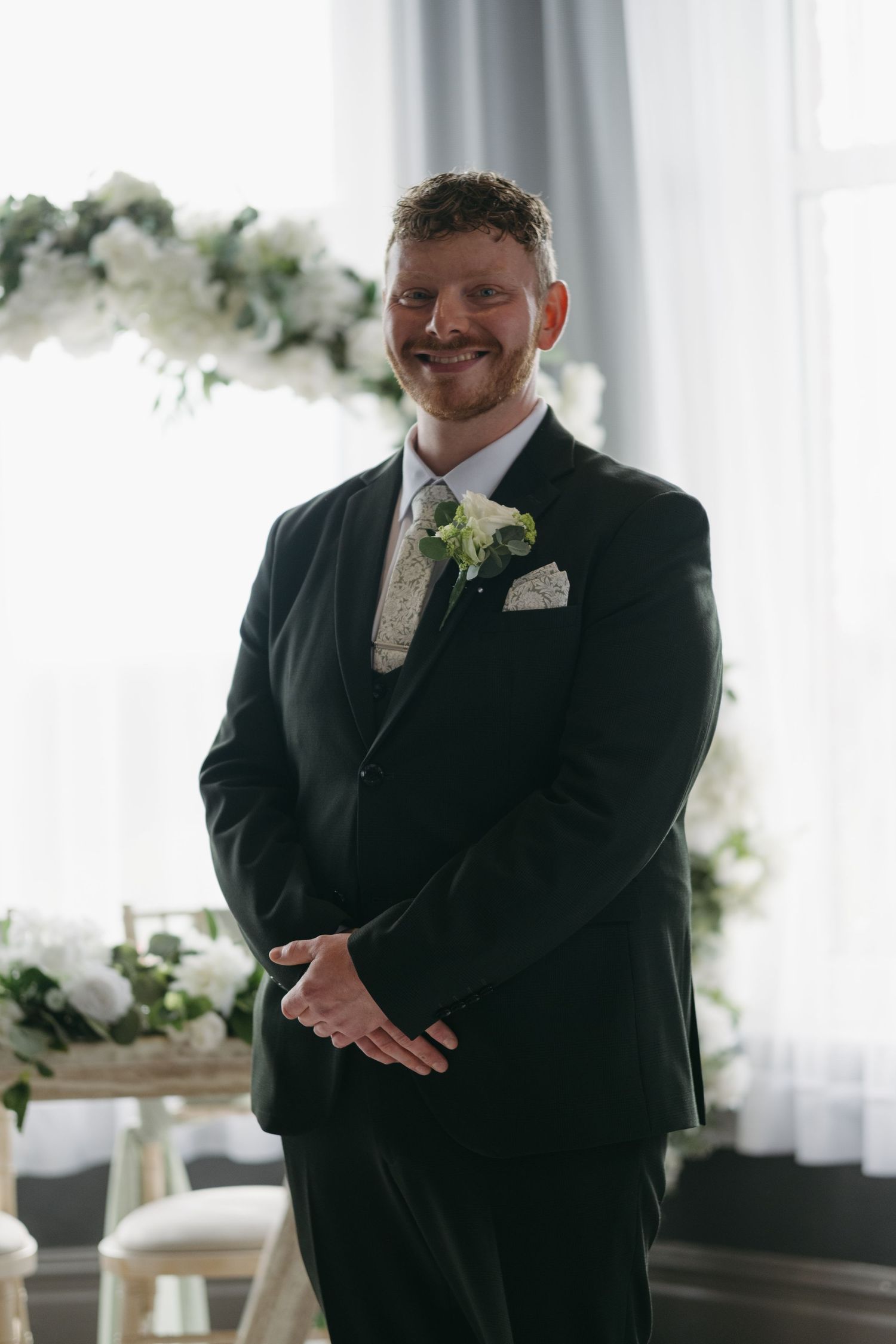 Smiling groom in dark suit with white boutonniere stands at wedding altar decorated with white flowers.