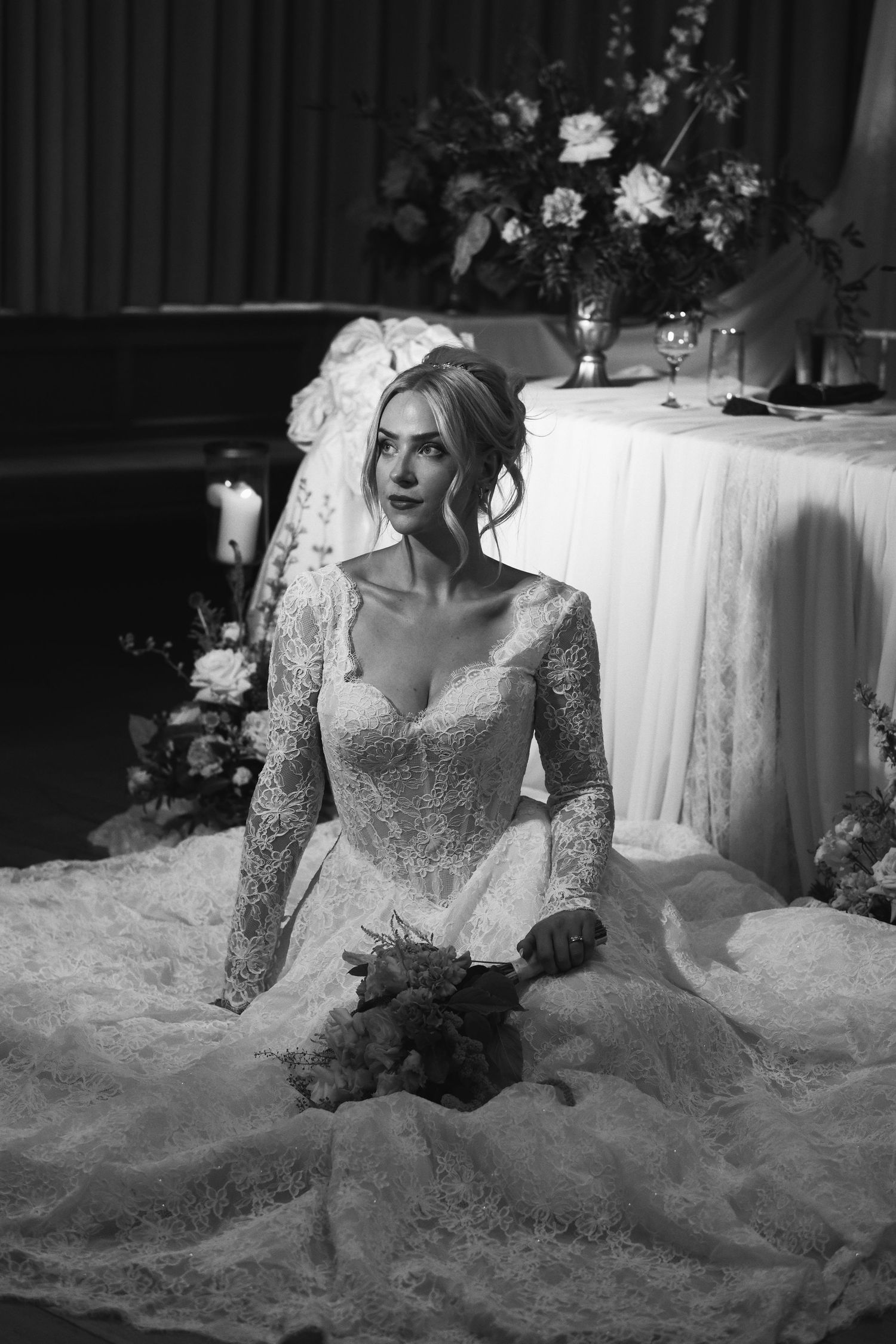 Bride in lace wedding gown sits gracefully on floor holding bouquet, black and white photo with floral backdrop.