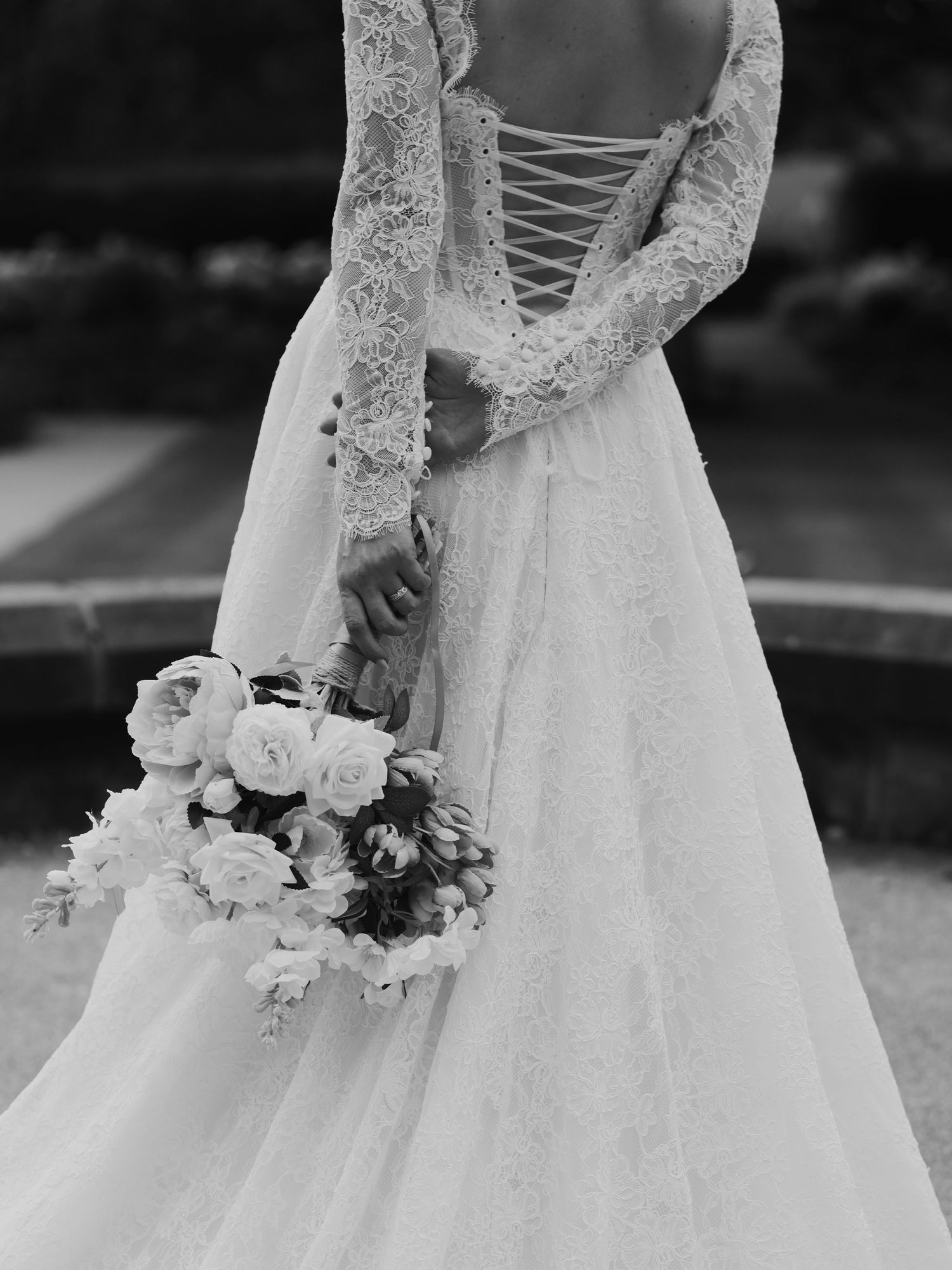 Black and white photo of a bride in a lace long-sleeve gown with corset back, holding a bouquet of roses.
