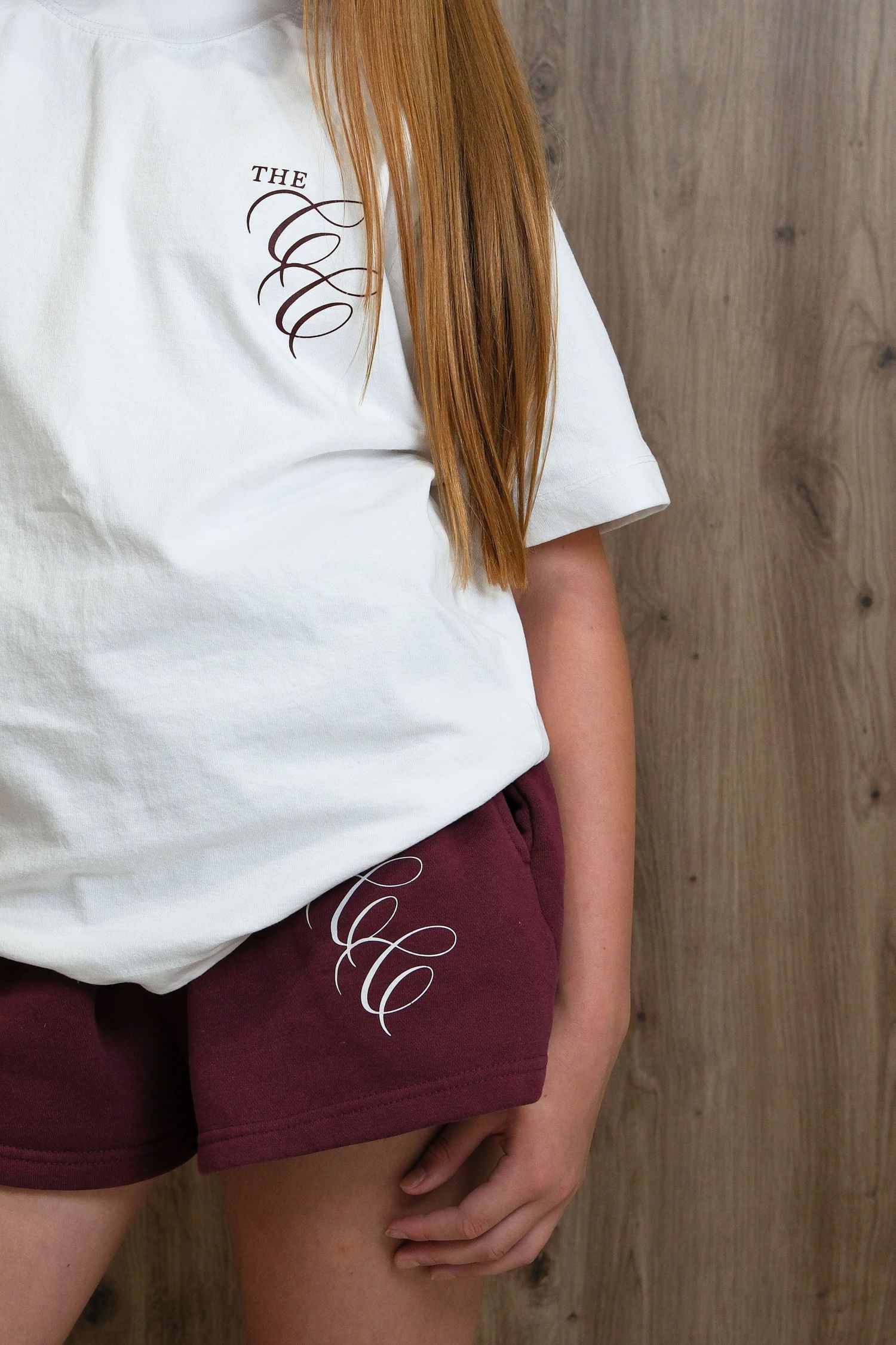 White t-shirt and burgundy shorts set with matching cursive CCC logo embroidery, shown on model against wood background.