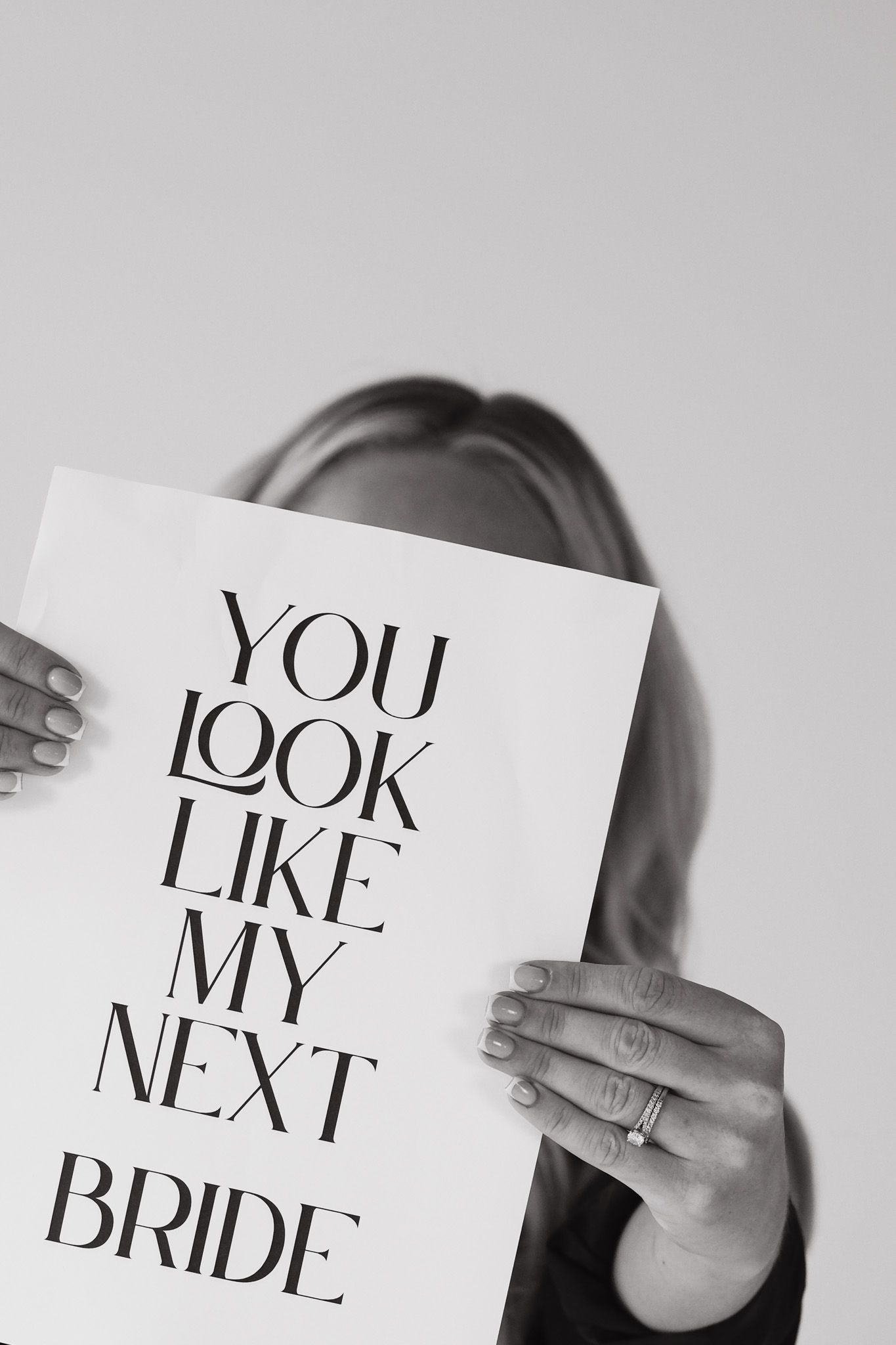 Black and white photo of woman holding sign reading You Look Like My Next Bride, covering her face.
