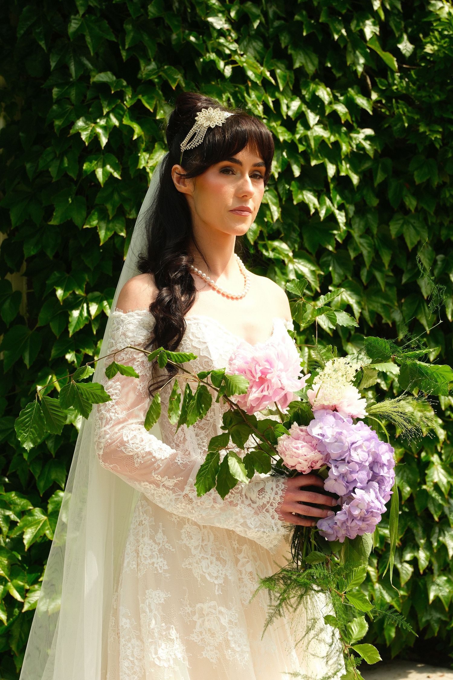 Elegant bride with dark hair in lace gown holding large pink and purple floral bouquet outdoors.