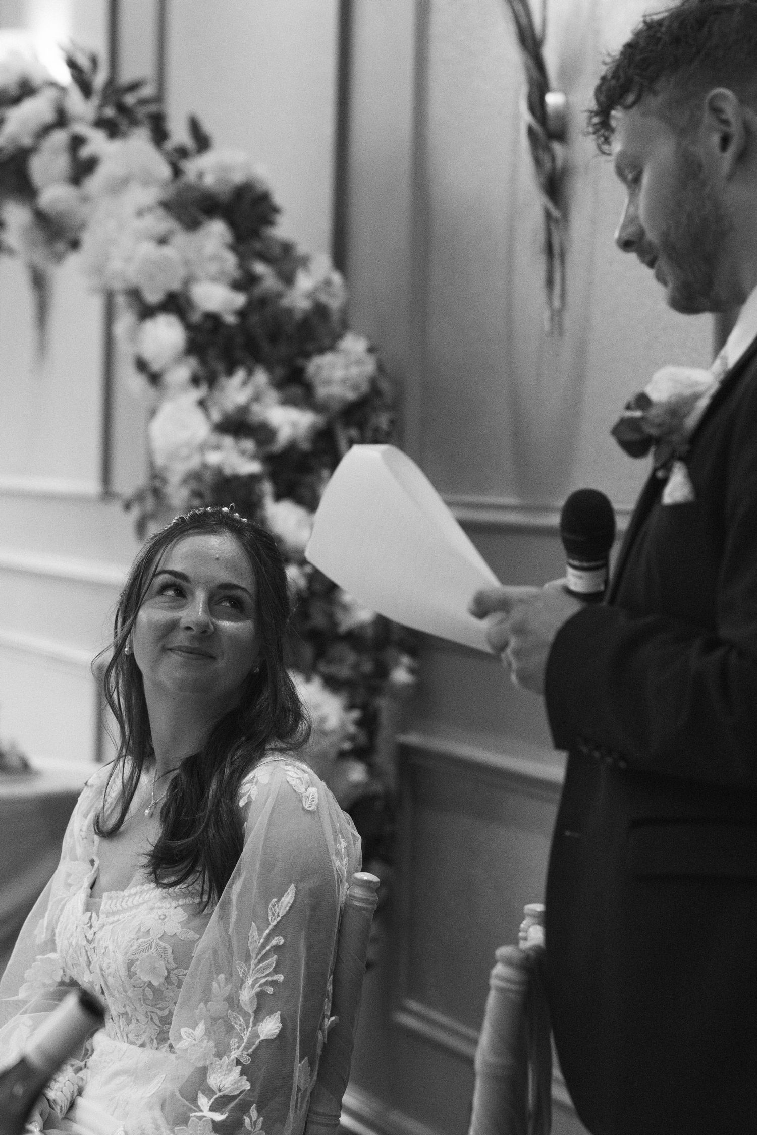Black and white wedding photo of bride gazing lovingly at groom reading vows, floral arrangement in background.
