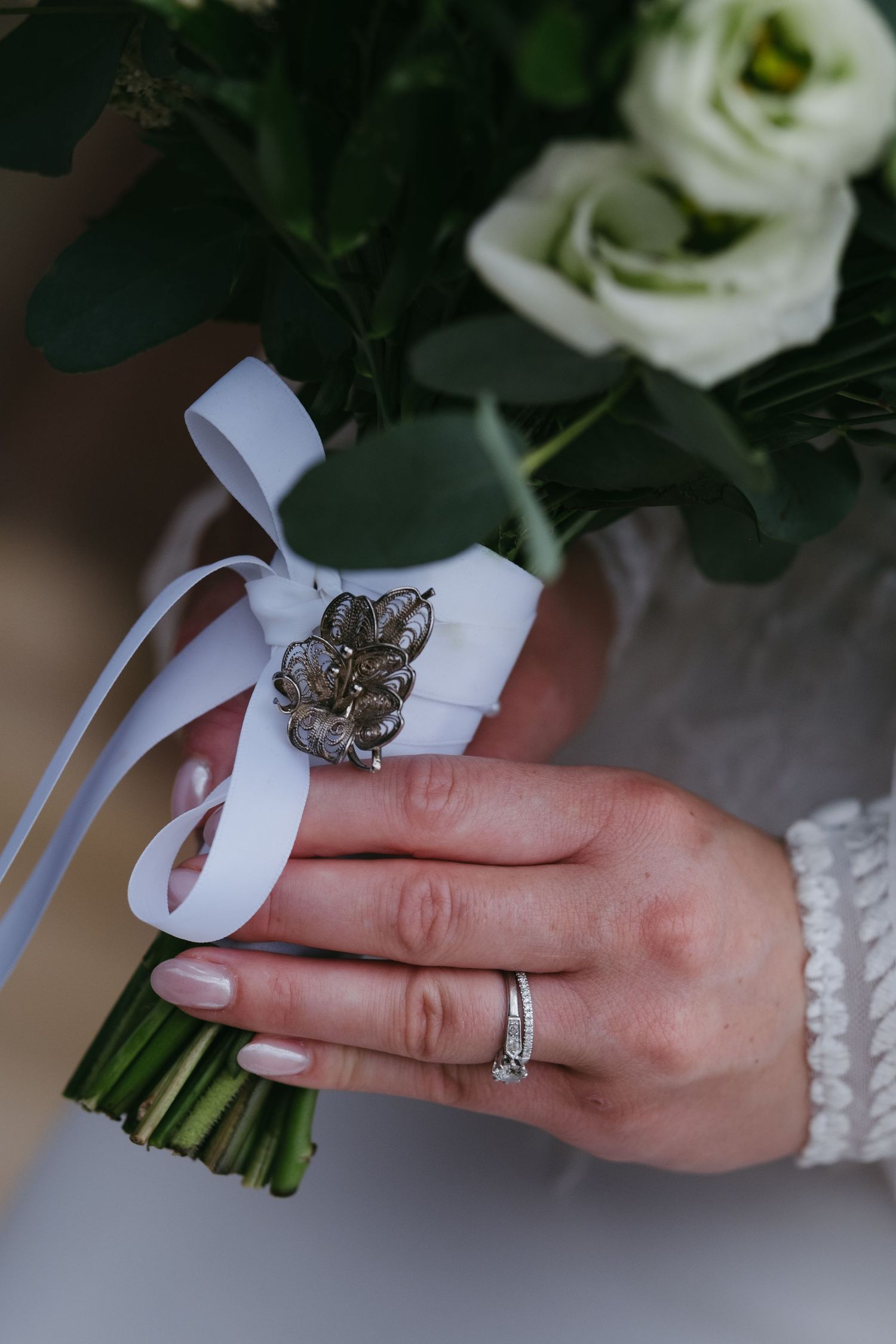 Bride holding white rose bouquet with silver brooch and ribbon, wearing diamond wedding ring and lace sleeve.
