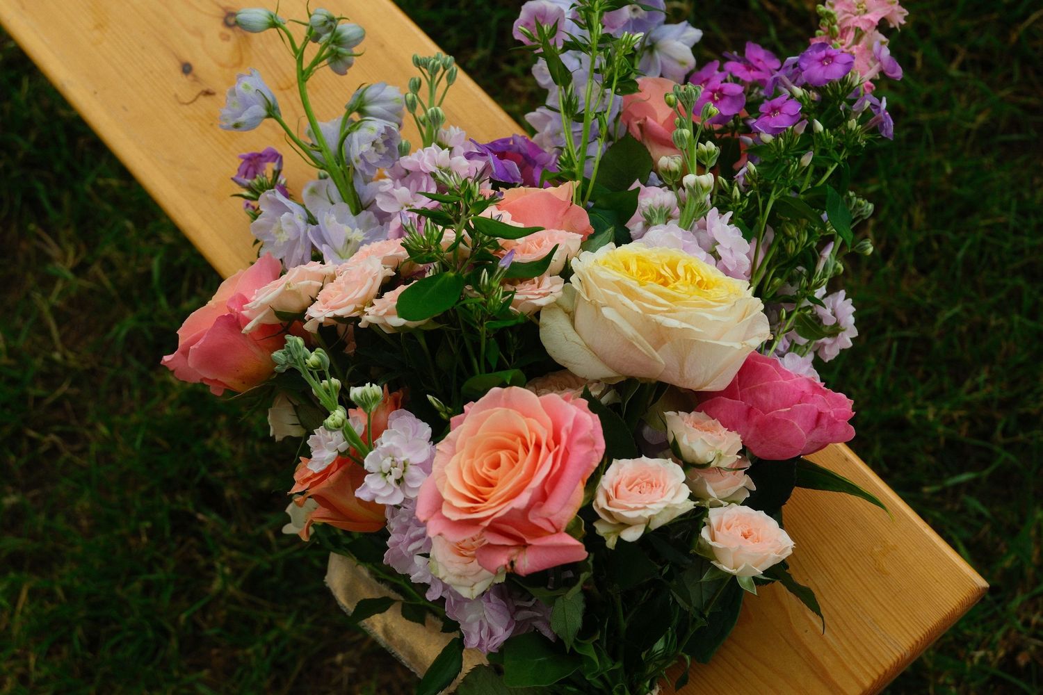 Colorful wedding bouquet with pink, peach, and white roses on a wooden bench outdoors.