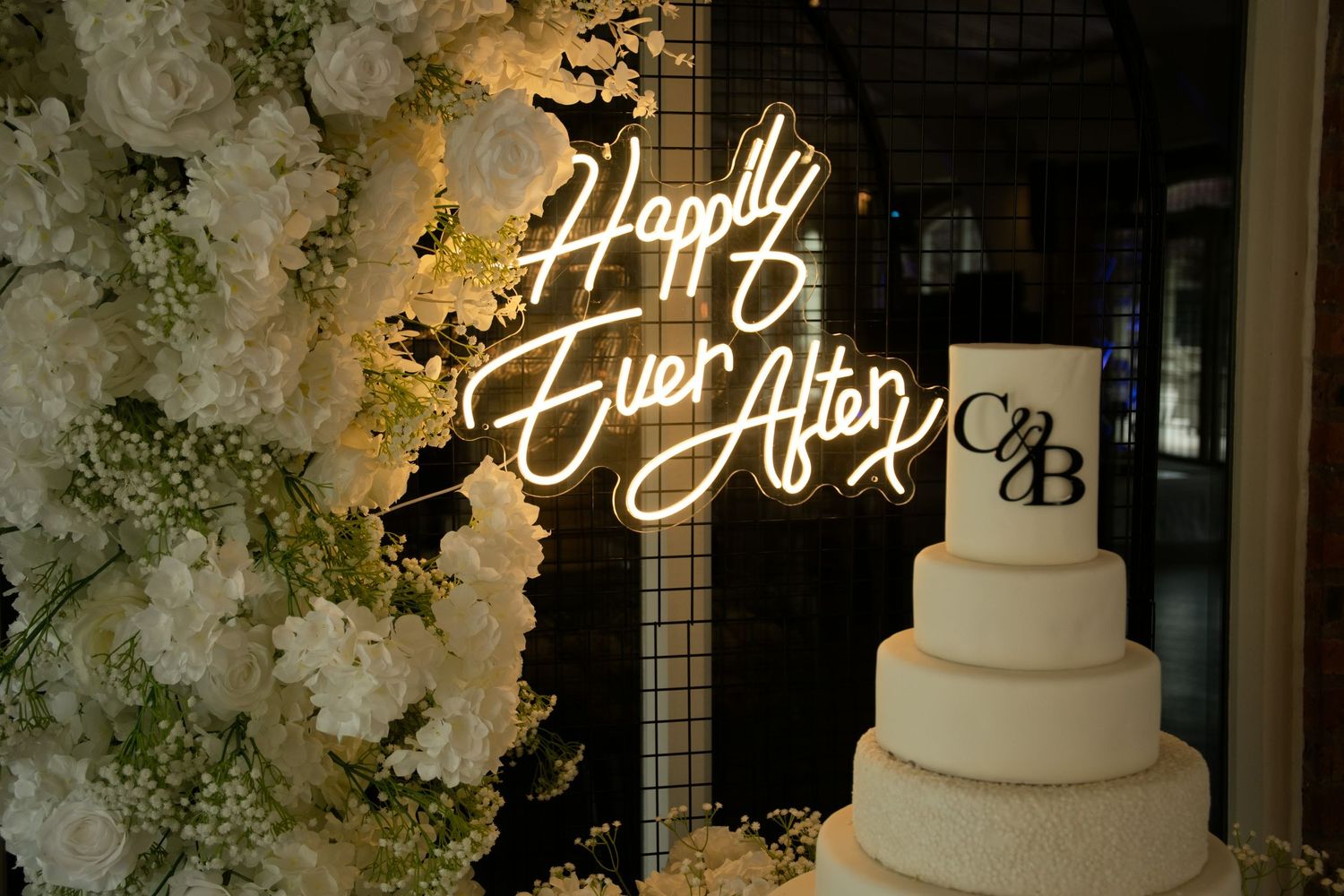 Elegant four-tier white wedding cake beside glowing Happily Ever After neon sign with floral backdrop.