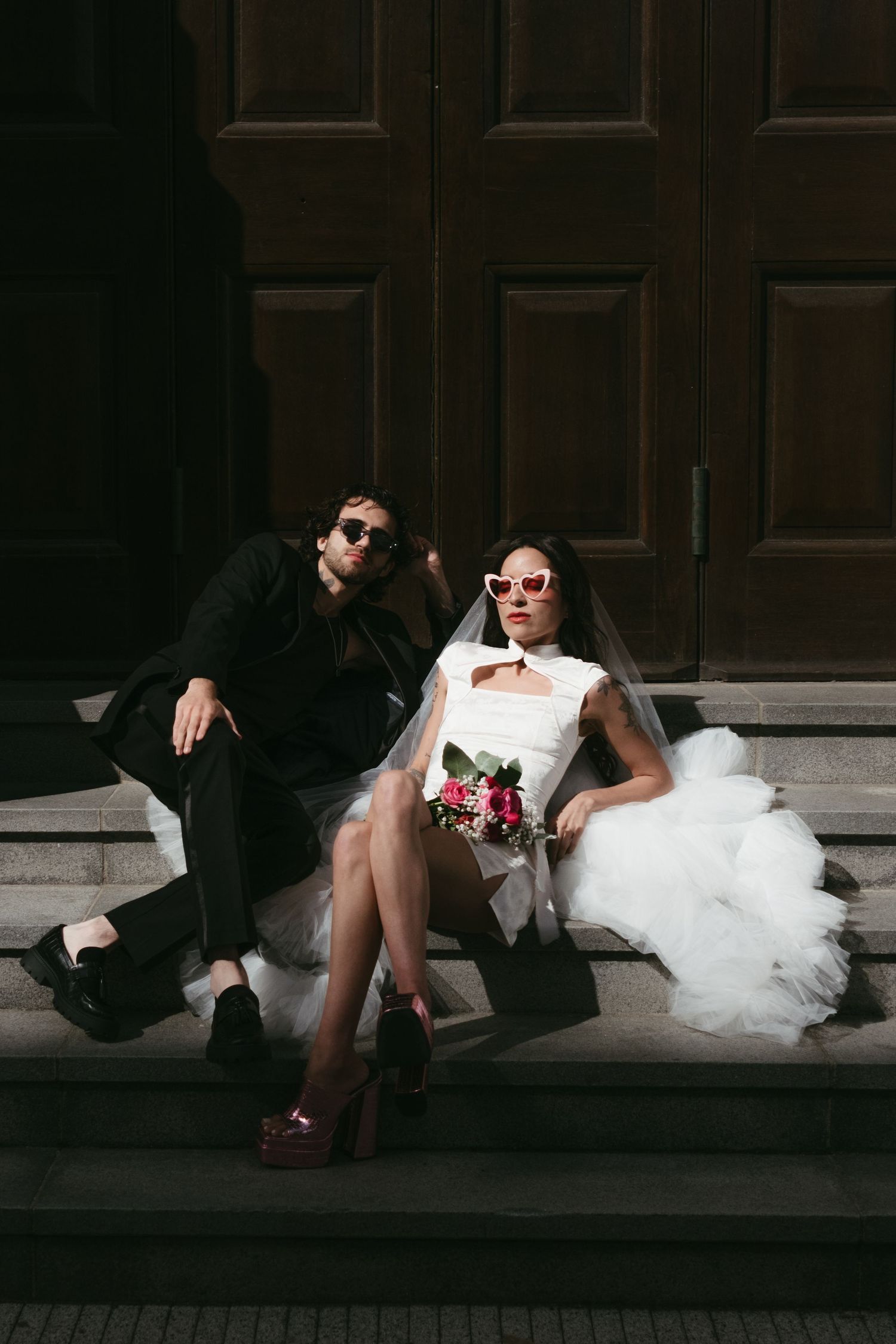 A stylish bride and groom in sunglasses sit on stone steps before large wooden doors, she holds a bouquet in a white gown.