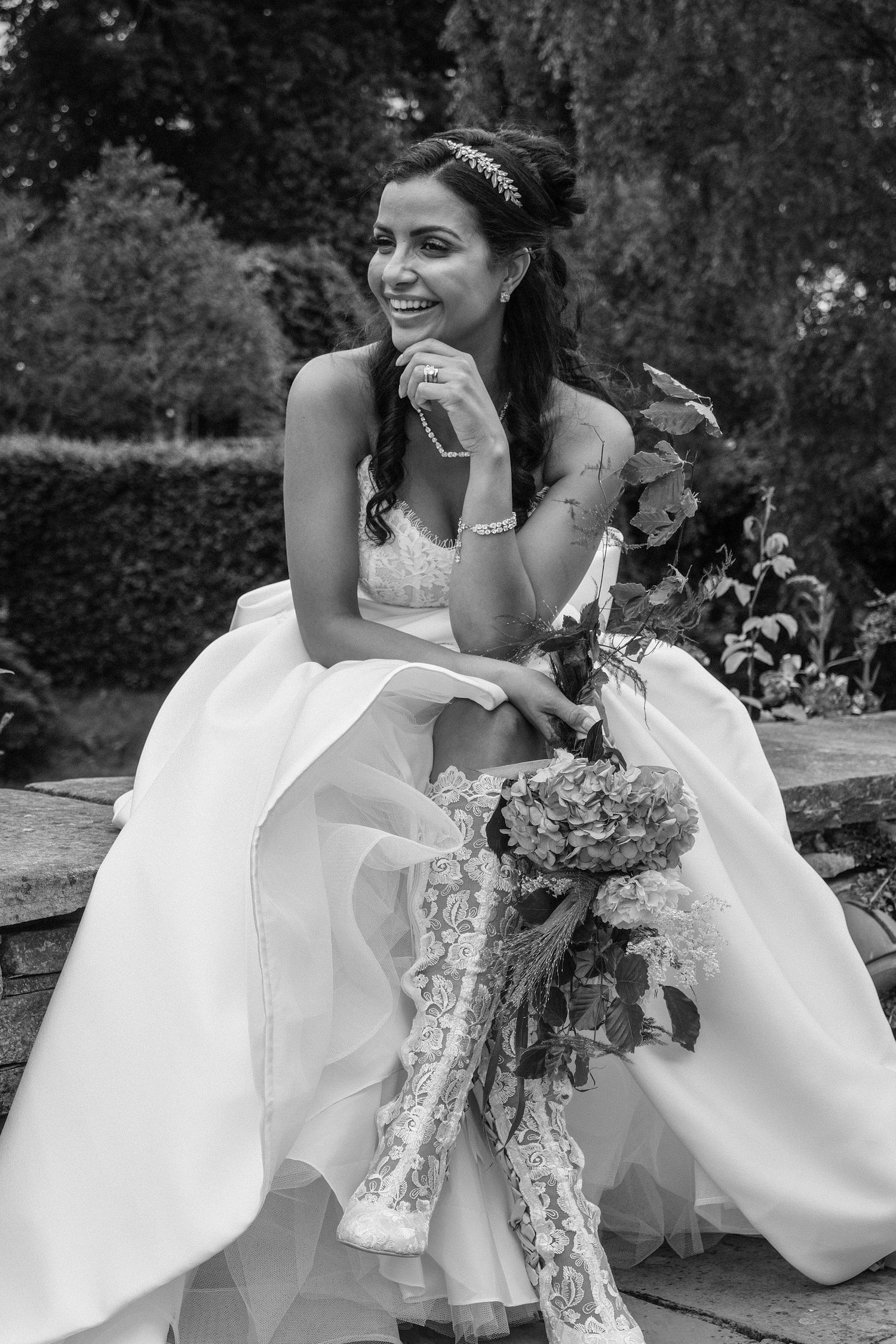 Black and white photo of a smiling bride in a white gown holding a bouquet, seated outdoors in a garden setting.