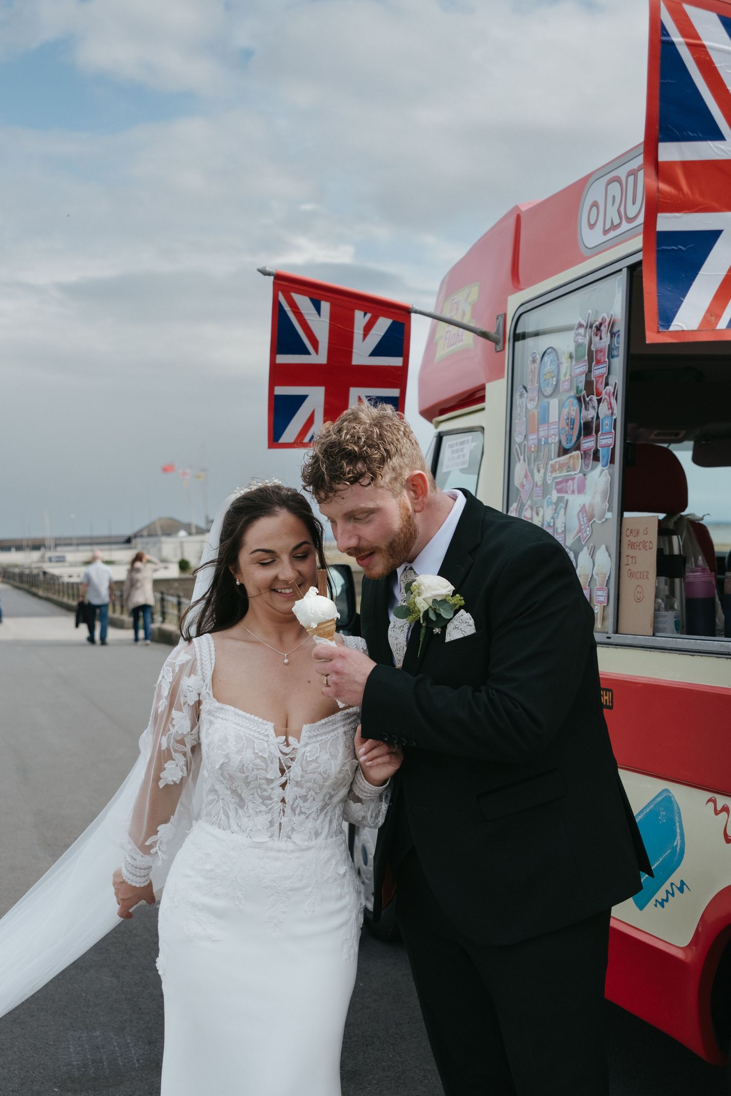 Bride and groom sharing an ice cream cone by a red British ice cream van with Union Jack flags on their wedding day.