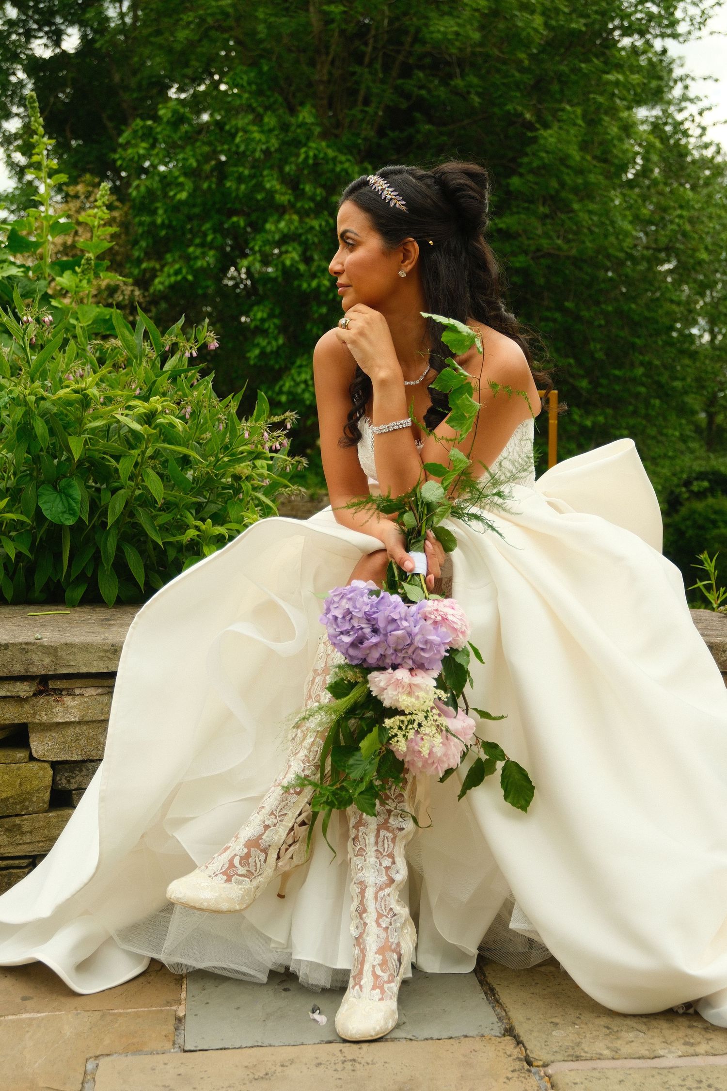 Bride in strapless ball gown with lace boots holding purple hydrangea bouquet seated on stone wall.
