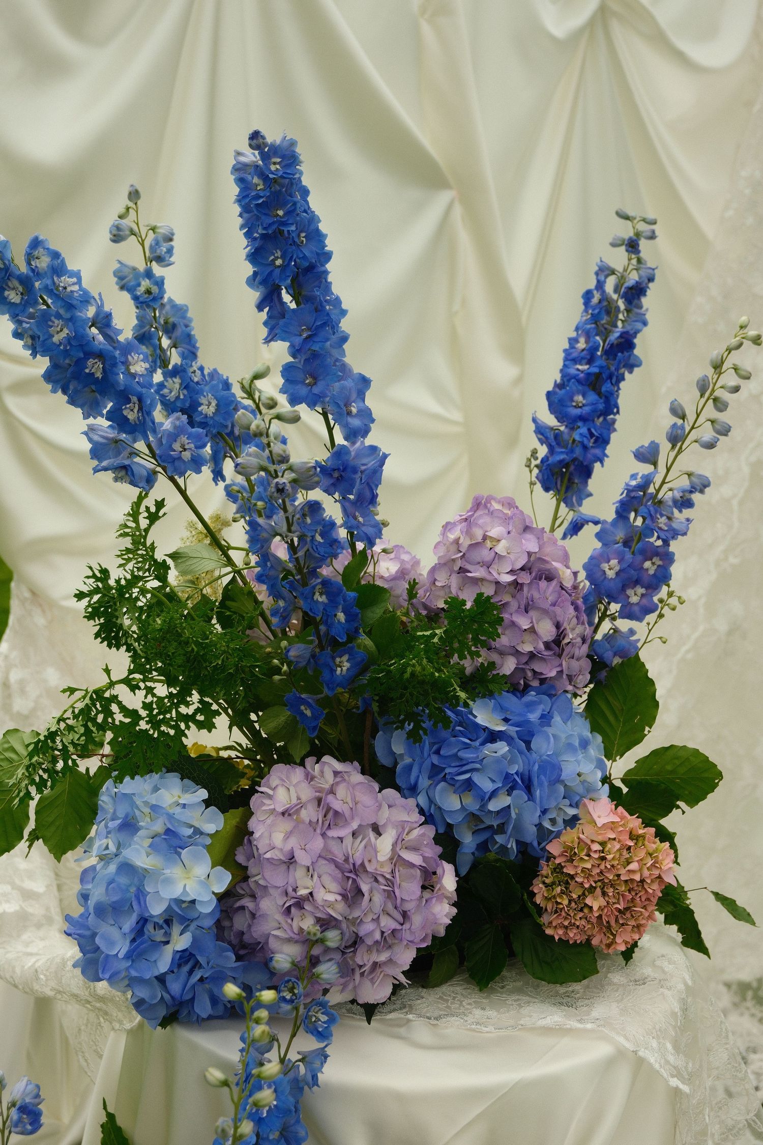 Wedding floral arrangement featuring blue delphiniums and purple hydrangeas against white draped fabric.