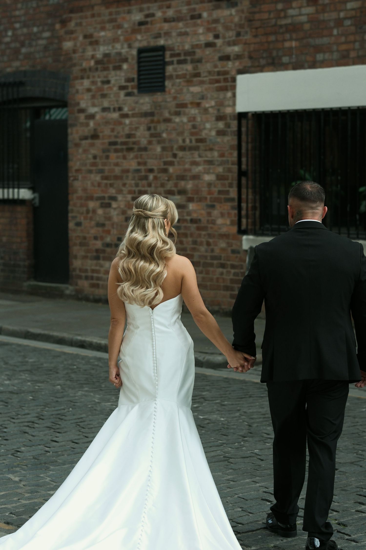 Bride and groom walking away hand in hand on cobblestone street, bride in fitted white gown with button back.