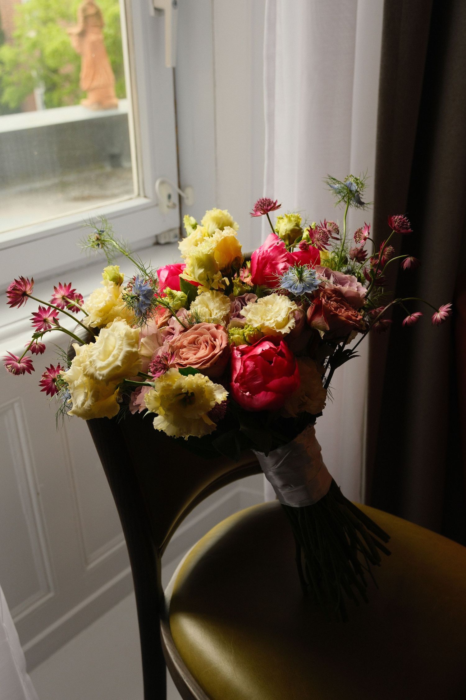Colorful wedding bouquet with pink peonies, yellow roses, and wildflowers resting on a chair by a sunlit window.