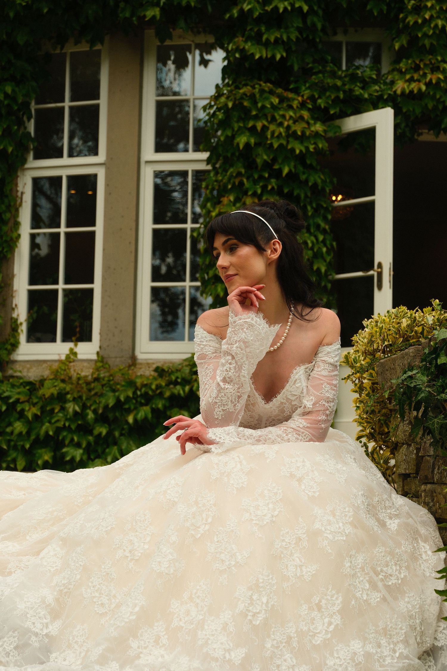 Elegant bride in off-shoulder lace wedding gown with pearl necklace seated outside ivy-covered manor house.