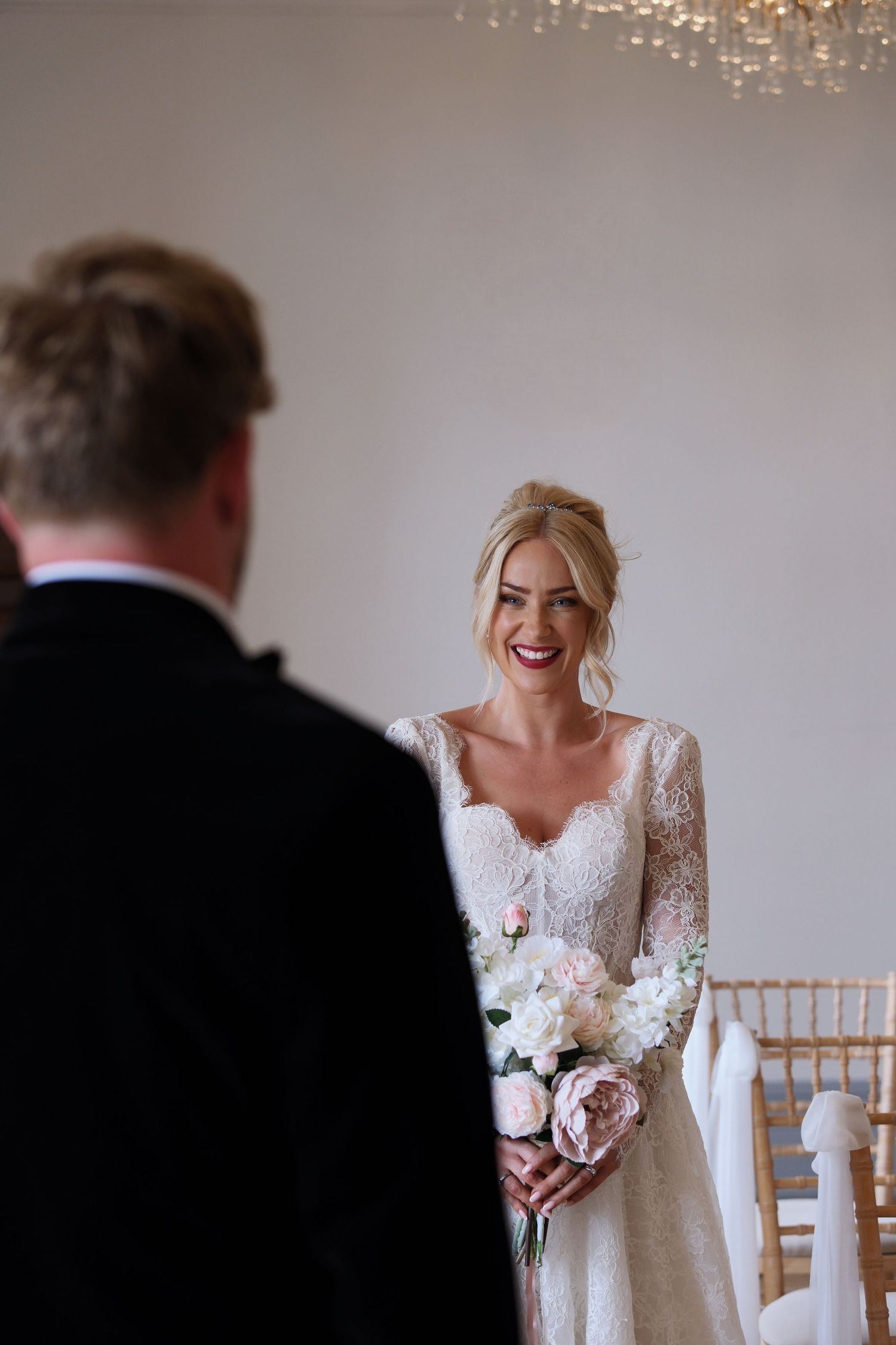 Smiling bride in lace long-sleeve gown holds pink and white bouquet facing groom during indoor wedding ceremony.