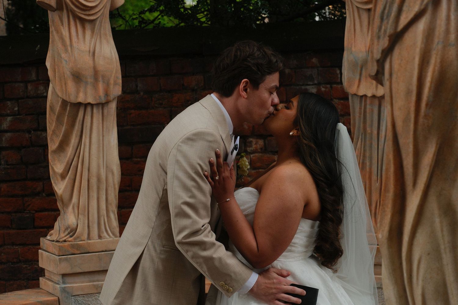 Bride and groom share their first kiss surrounded by classical stone statues at an outdoor wedding ceremony.