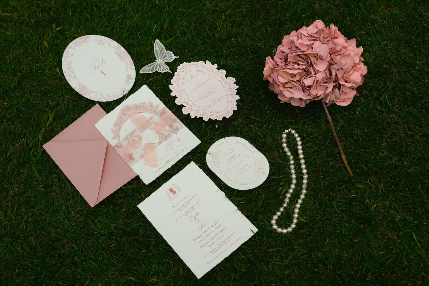 Flat lay of pink wedding stationery suite with pearl necklace and dried hydrangea on green grass.
