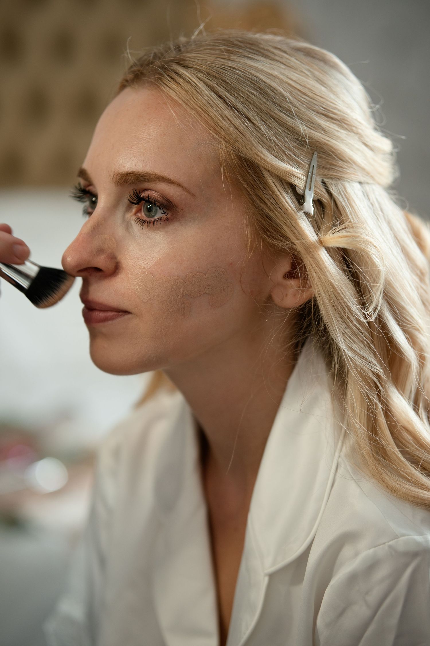Blonde bride getting makeup applied with a brush on her nose during bridal preparation.