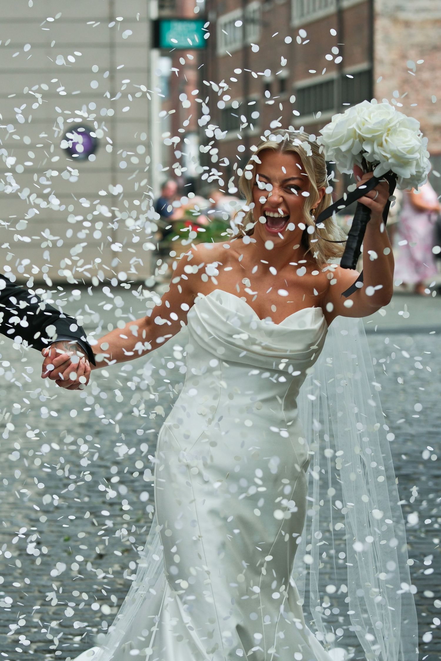 Joyful bride in white gown celebrating with confetti cannon on city street.